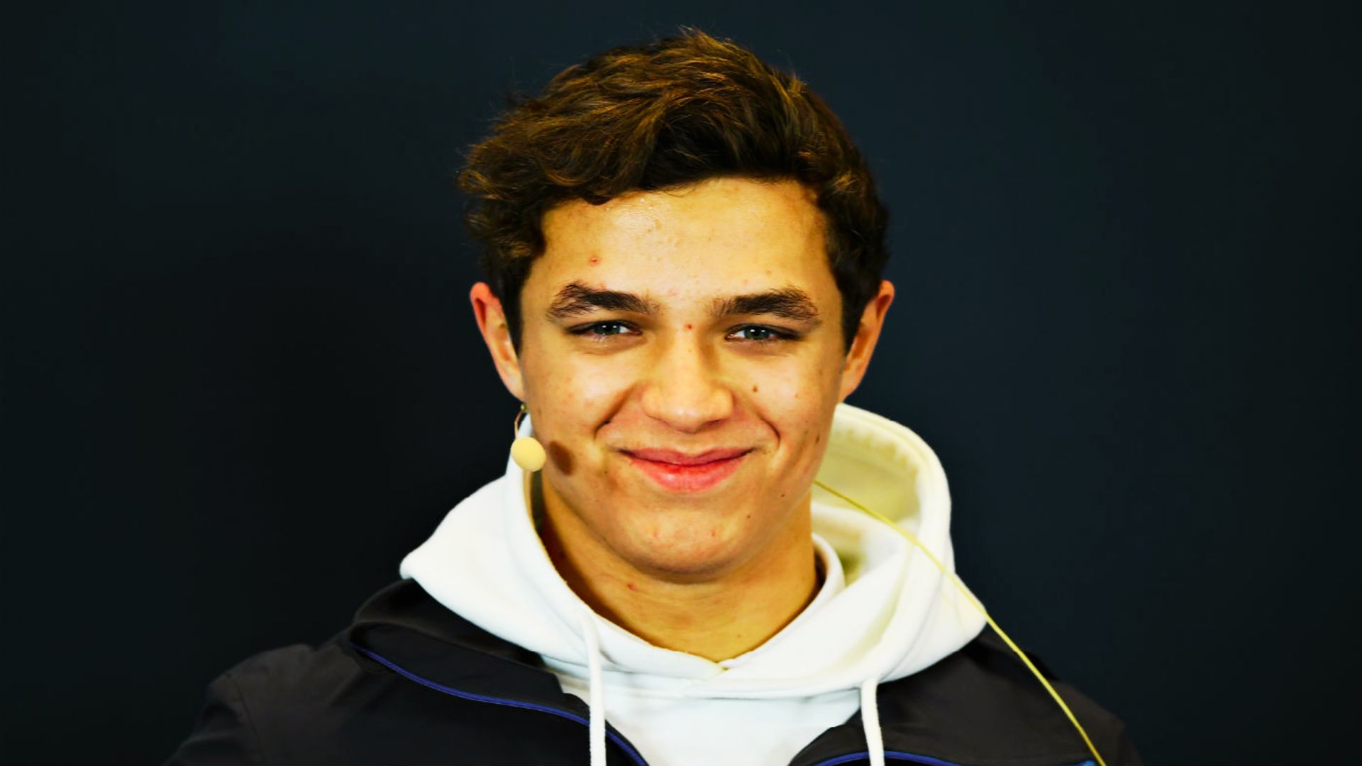 Lando Norris smiles in a white hoodie, wearing a headset mic.