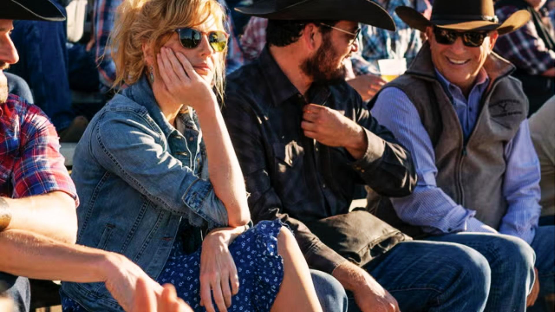 Beth Dutton sits at rodeo in denim jacket, sunglasses, and blue printed midi skirt among cowboy hats.