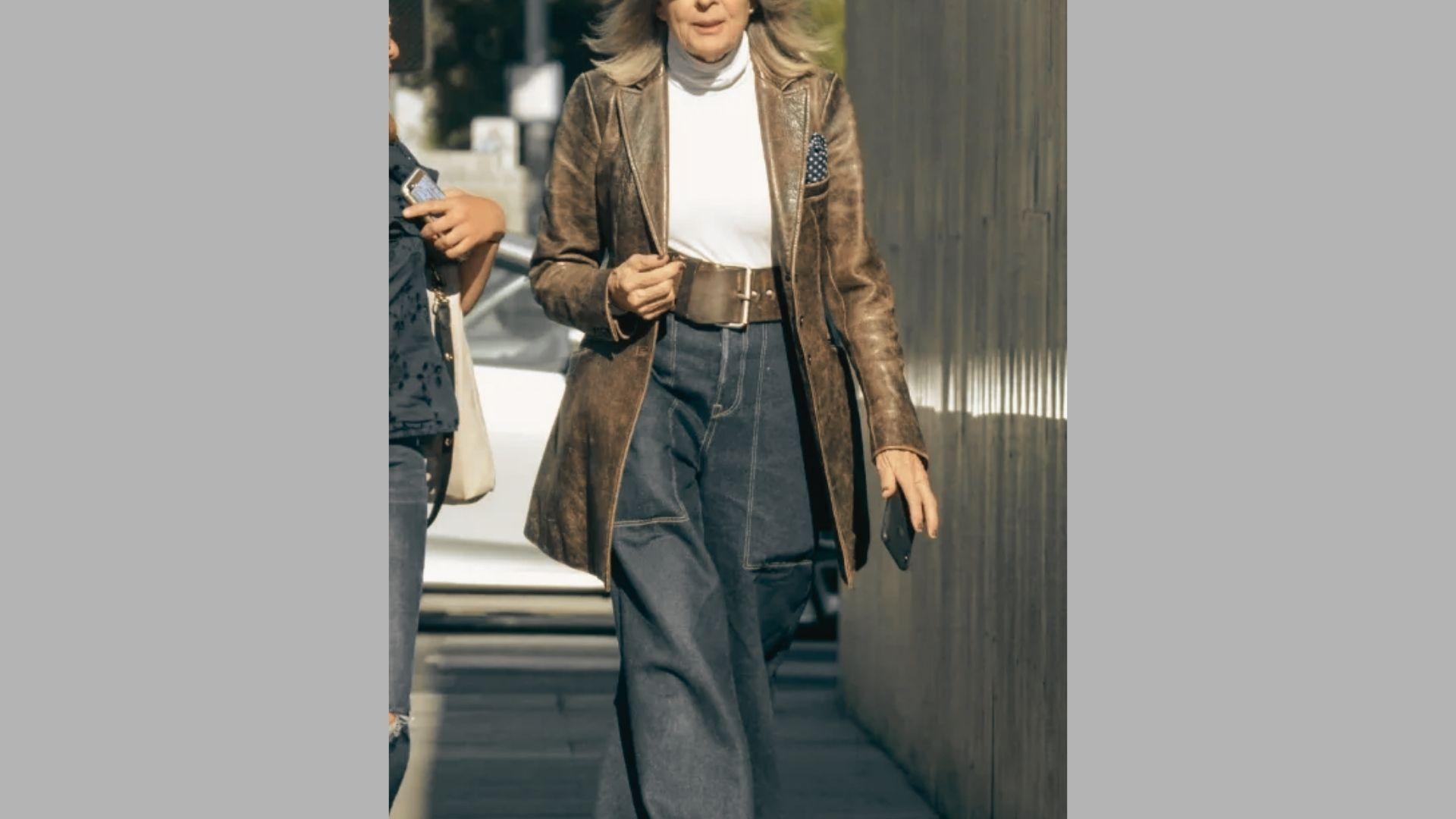 Woman walks outdoors in brown leather jacket, white turtleneck, and wide jeans holding phone