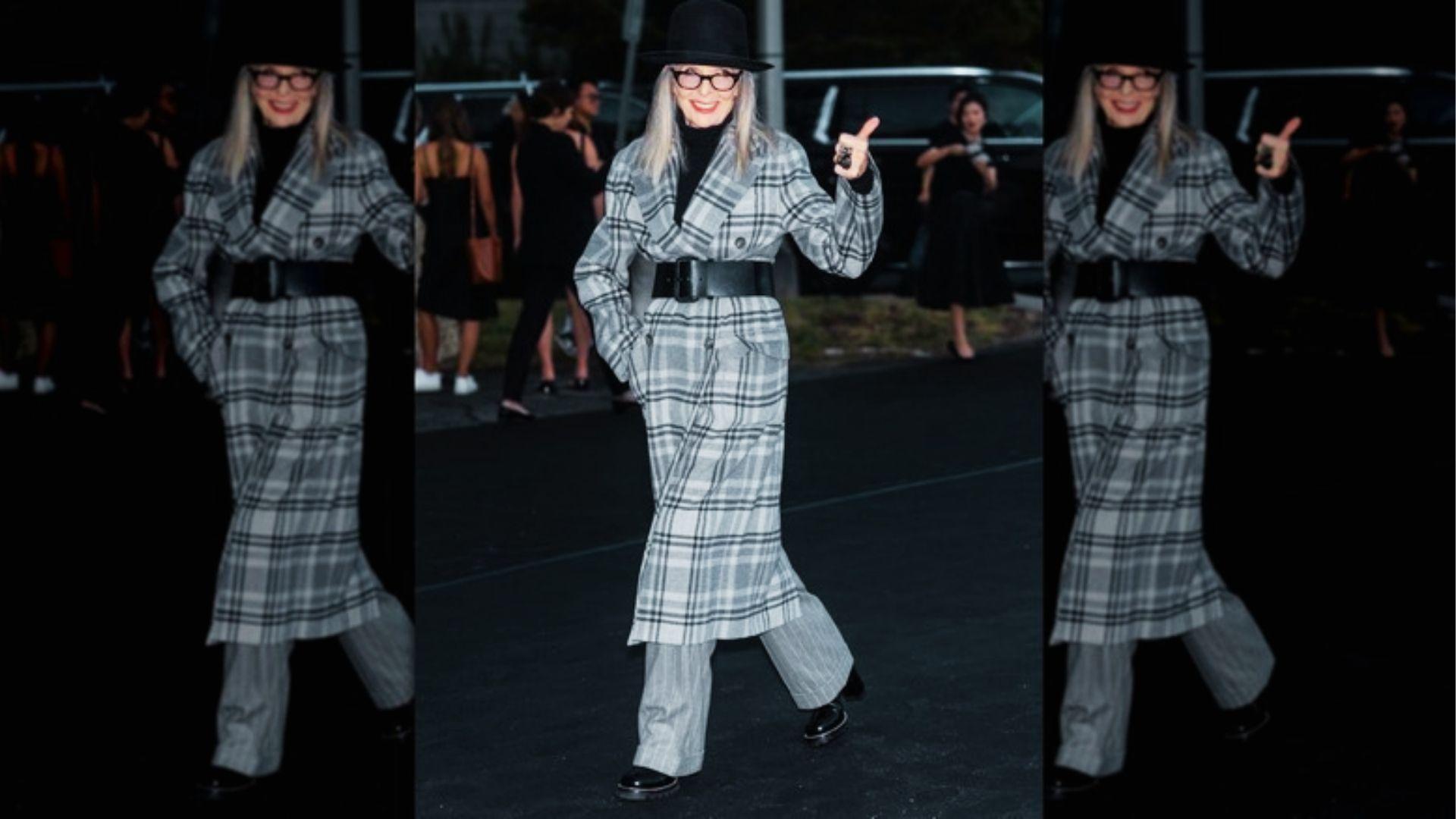 Woman in plaid belted coat, black turtleneck, and hat gestures while walking nighttime street