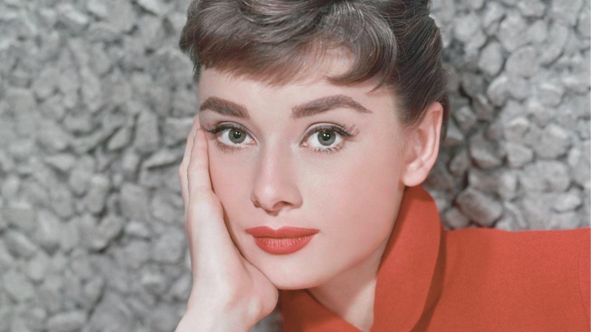 Color portrait of Audrey Hepburn in a vibrant red wool coat with a rounded collar, showcasing her classic pixie bangs and red lipstick.