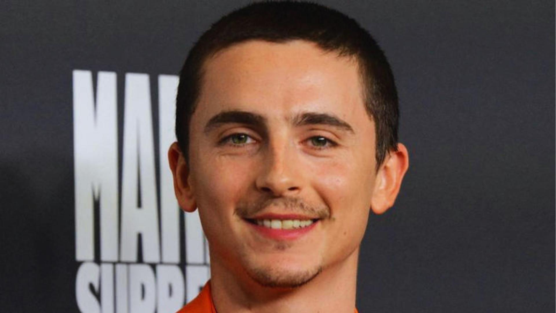 Timothée Chalamet debuts a short buzz cut and thin mustache in an orange jacket, a major departure from his signature curls.