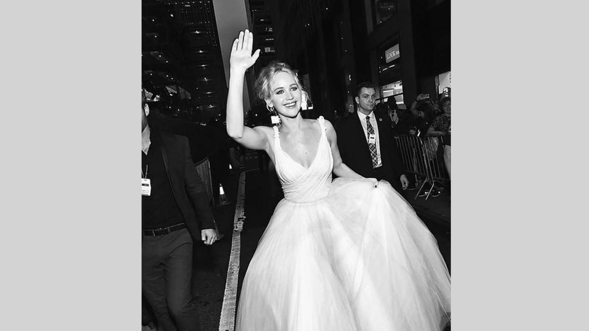 Black and white photo of actress waving in white tulle gown surrounded by security