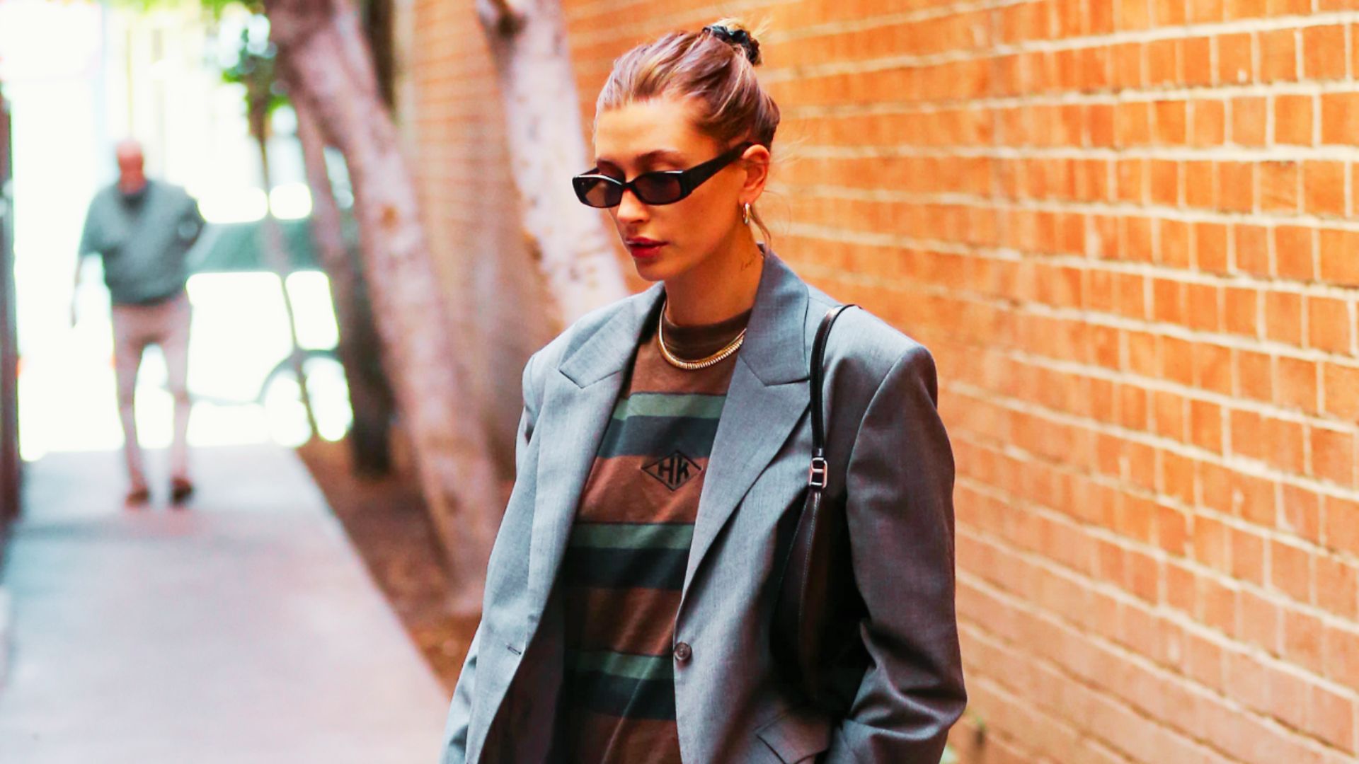 Hailey Bieber walks along a brick wall in an oversized gray blazer layered over a brown striped tee.