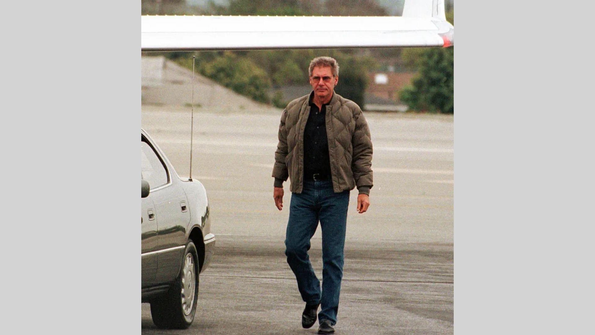 Harrison Ford walks airport tarmac in puffer jacket, black shirt, and jeans near plane