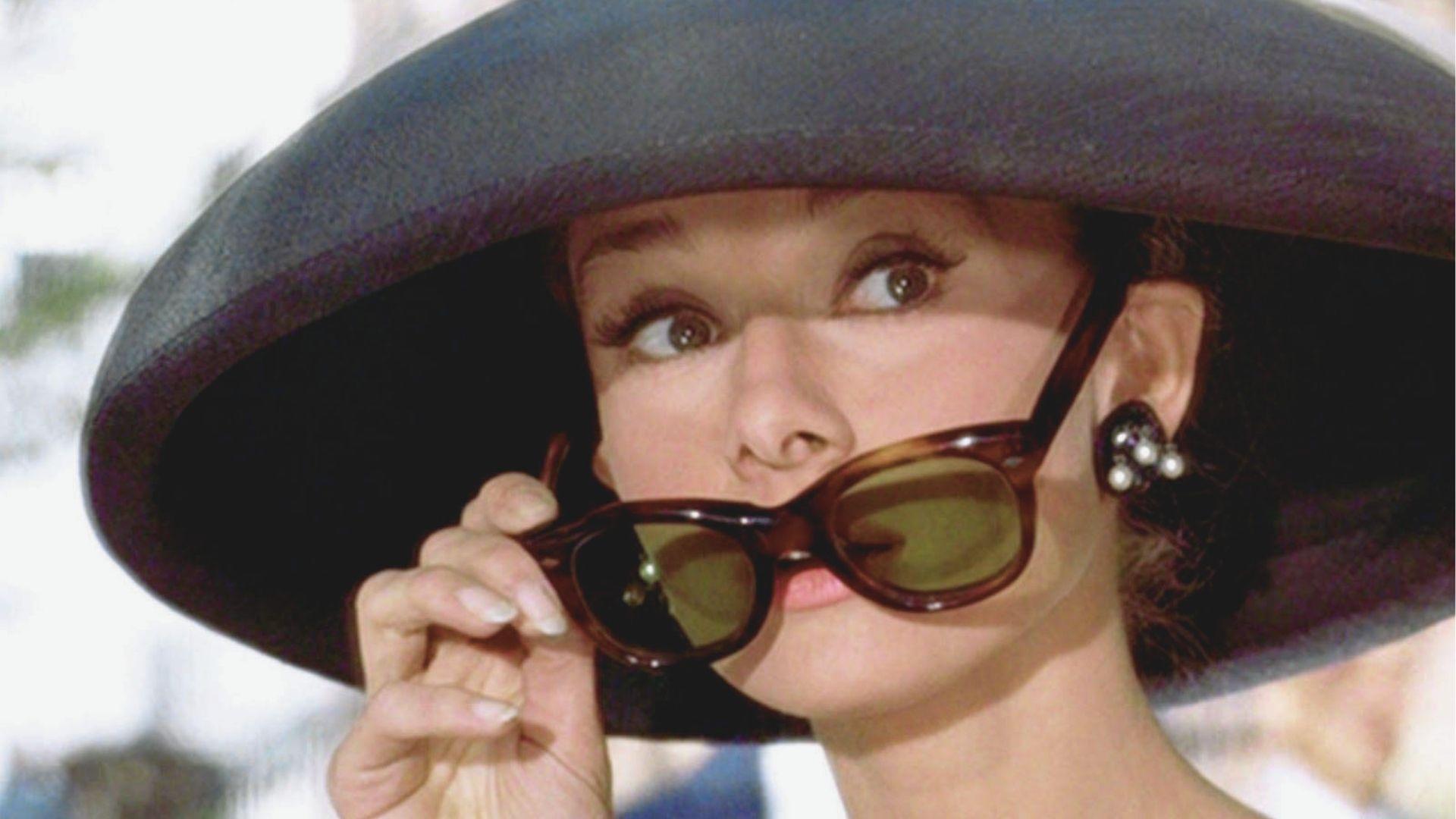 Audrey Hepburn as Holly Golightly peeks over oversized tortoiseshell sunglasses in a wide-brimmed black hat and pearl earrings.