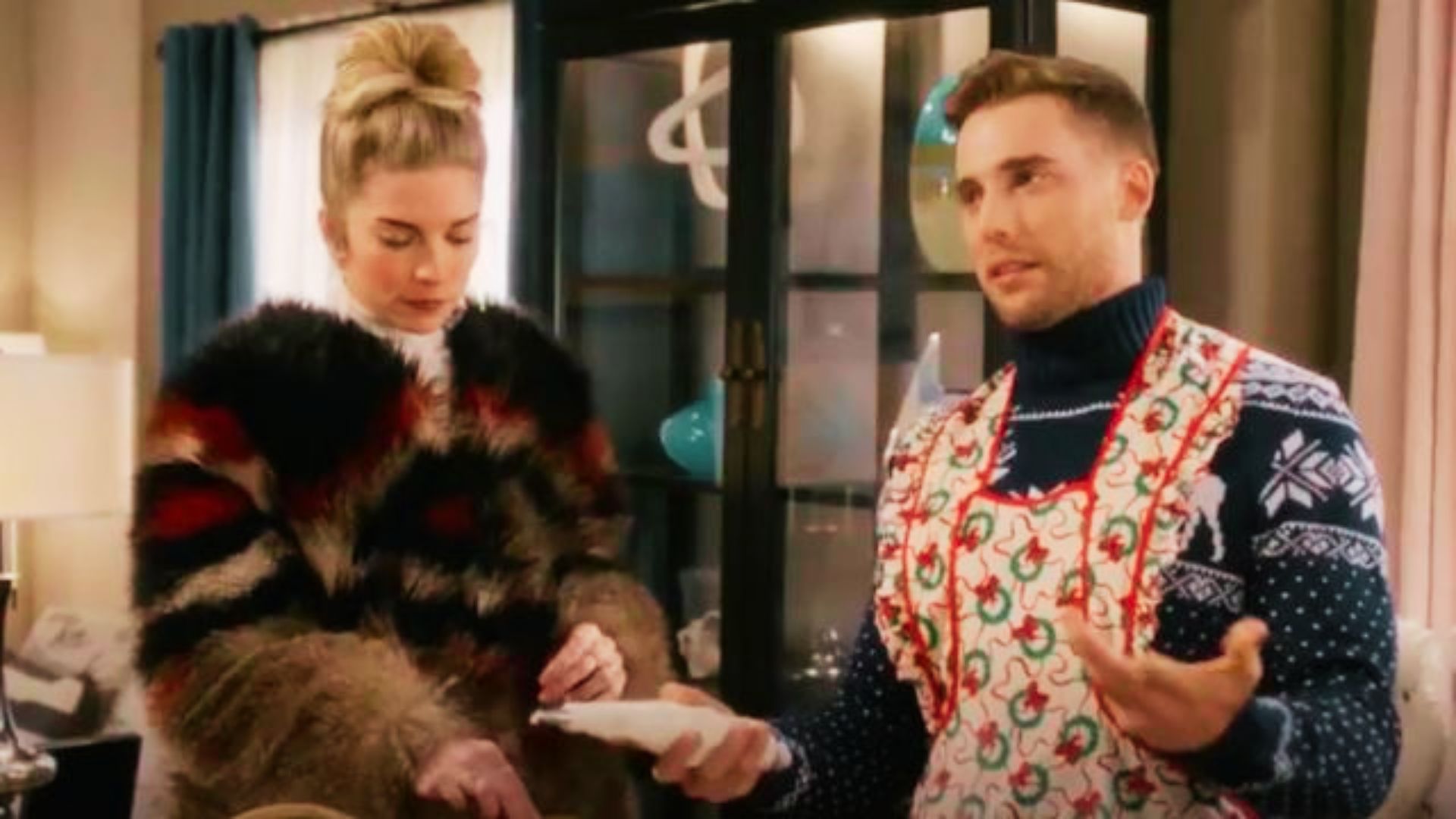 Alexis stands next to a man in a holiday apron, wearing an oversized striped faux fur coat.