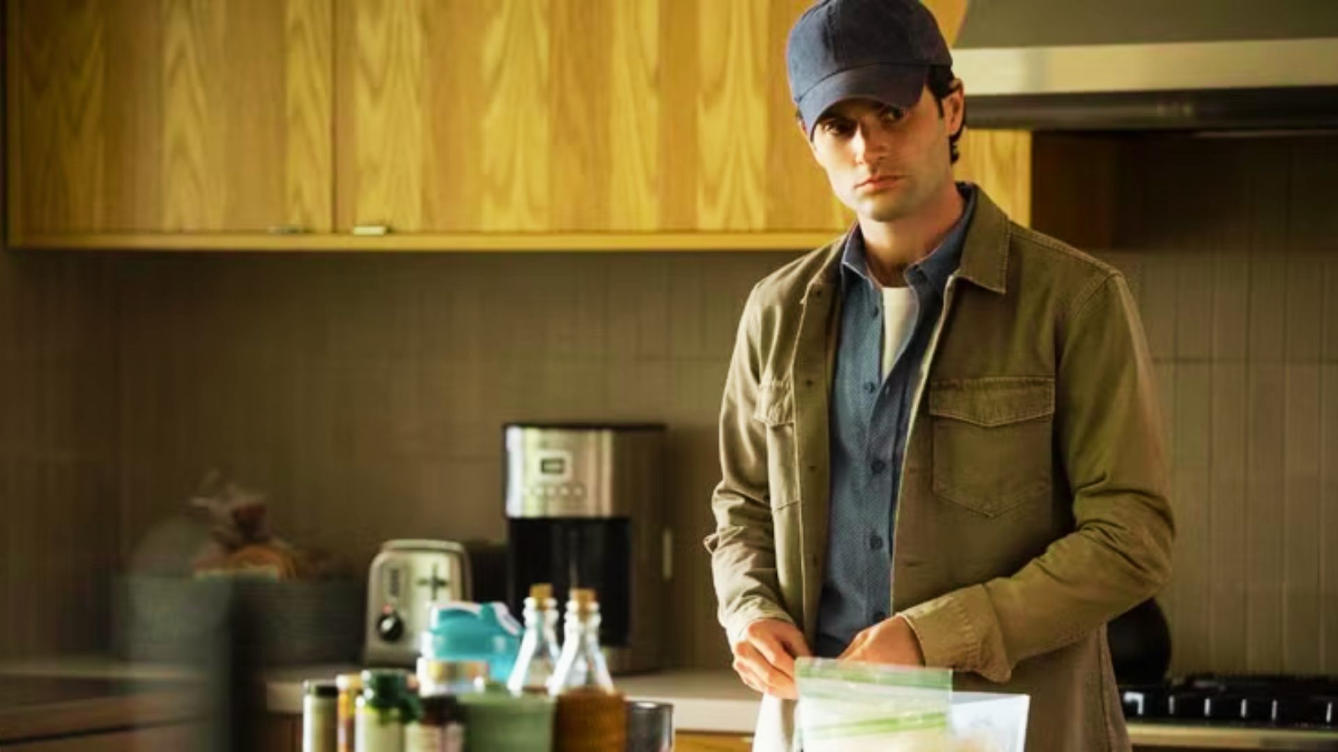 Joe Goldberg stands in a kitchen wearing a tan chore jacket, blue shirt, and navy cap.