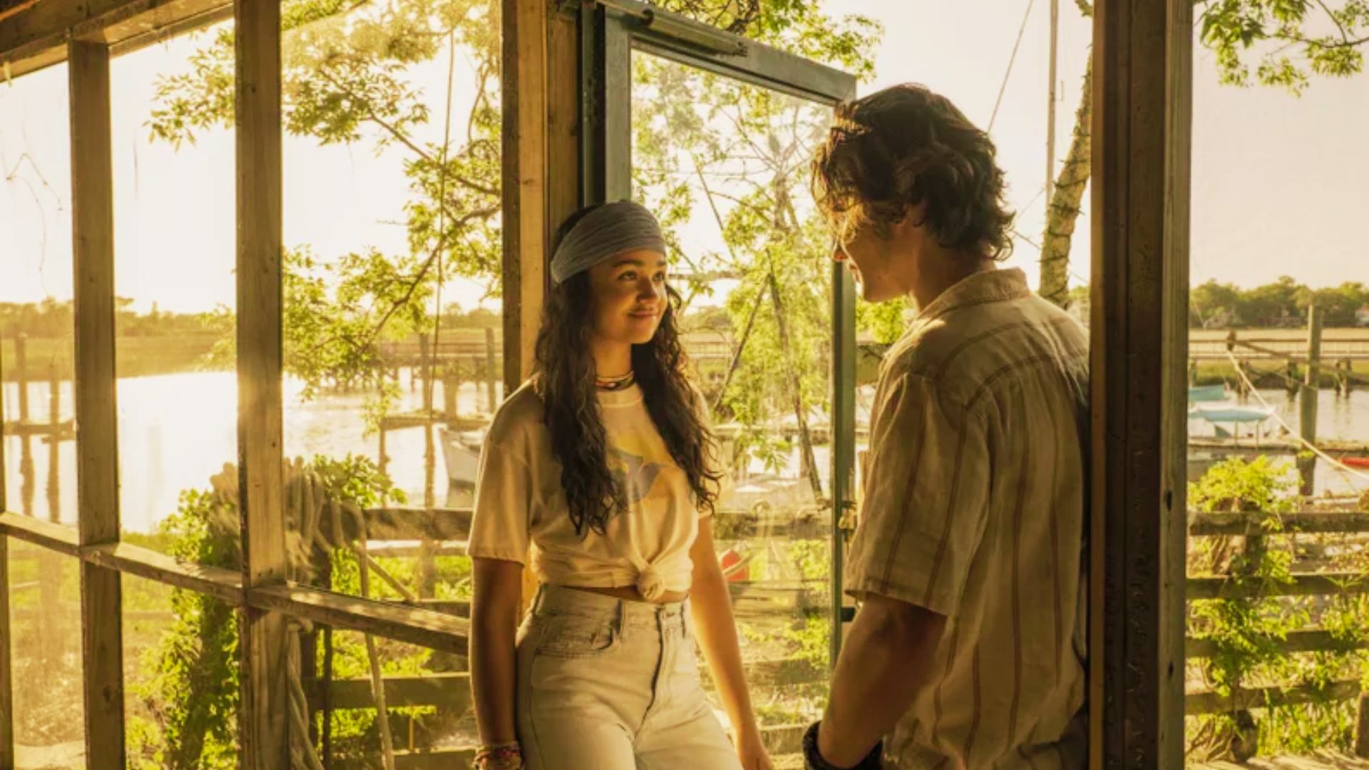 Kiara talks to John B in a sunlit beach shack wearing a knotted white graphic tee, light-wash jeans, and a grey headband.