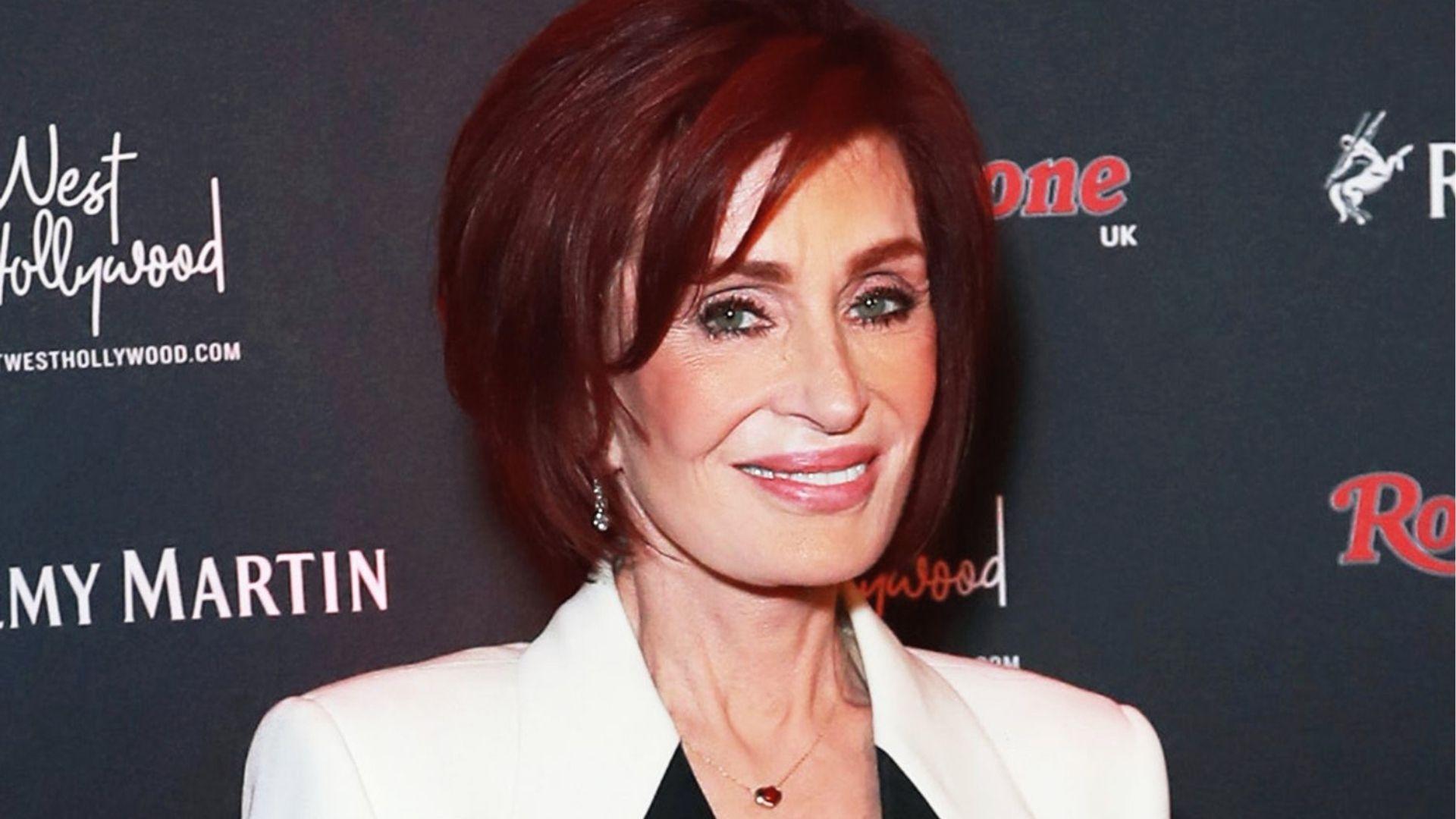 Sharon Osbourne smiles in a white blazer over black top with heart pendant necklace