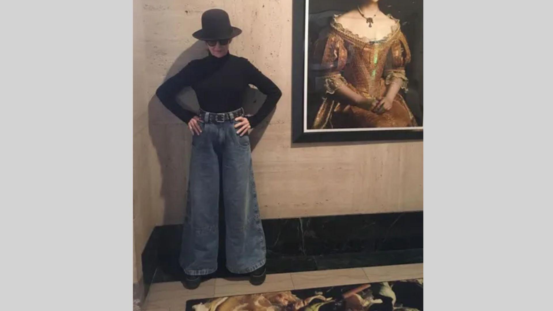 Woman in black turtleneck, wide jeans, and bowler hat poses by classic portrait painting