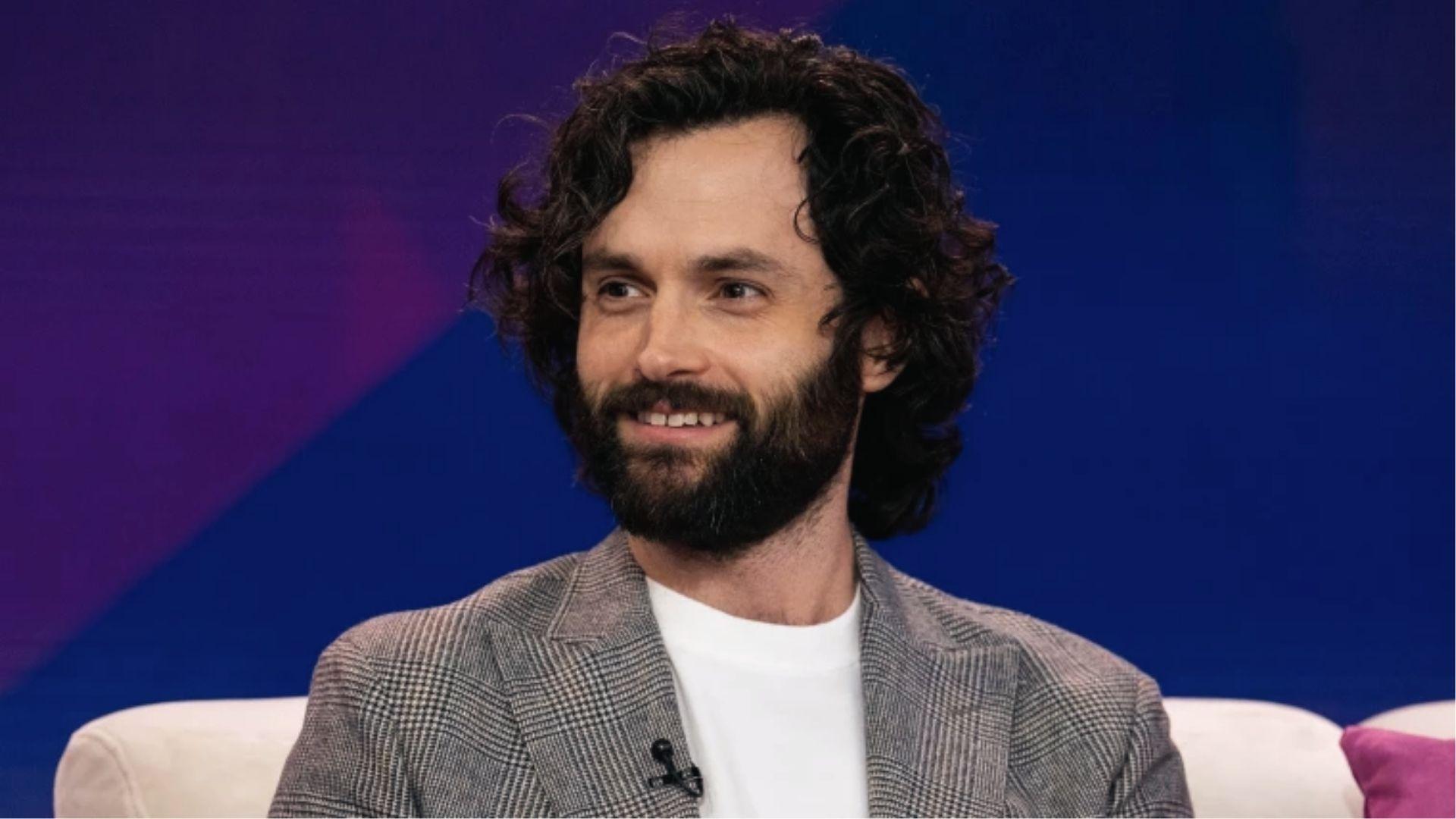 Penn Badgley smiles with long loose curls and full beard wearing plaid blazer on talk show set.
