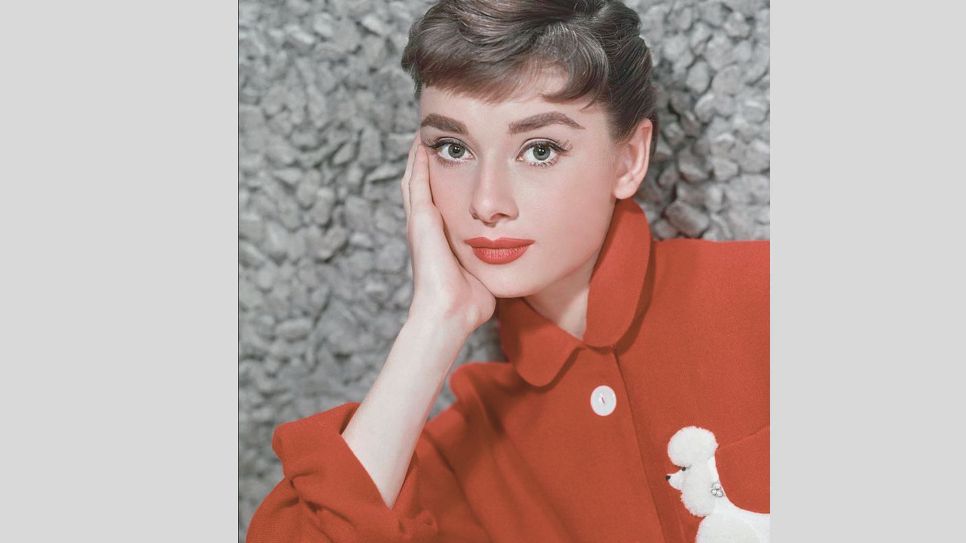 Close-up portrait of Audrey Hepburn resting her face on her hand, wearing a bold red collared coat with a white poodle appliqué.