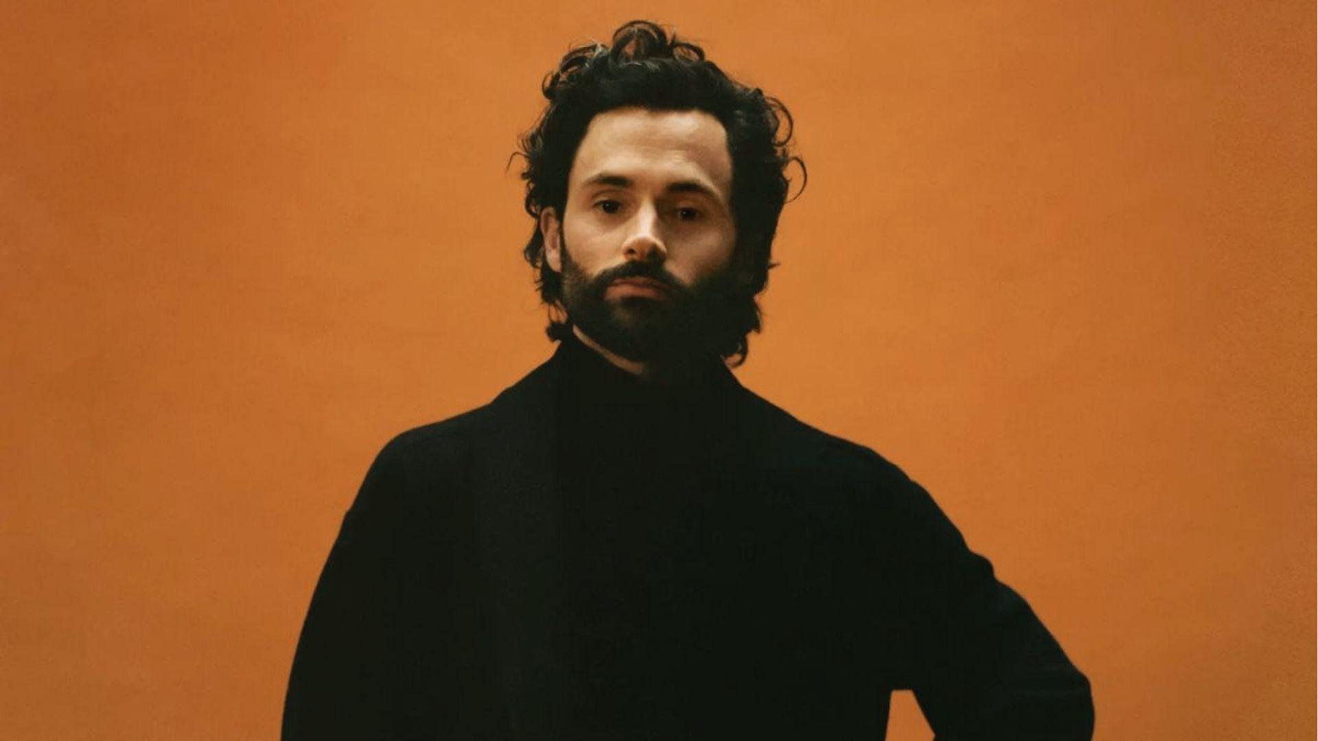 Penn Badgley wears longer wavy hair with natural texture against an orange backdrop portrait.