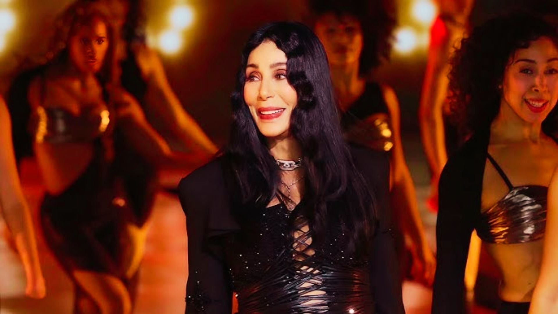 Cher onstage with dancers, wearing a black lace-up corset bodysuit covered in crystals.