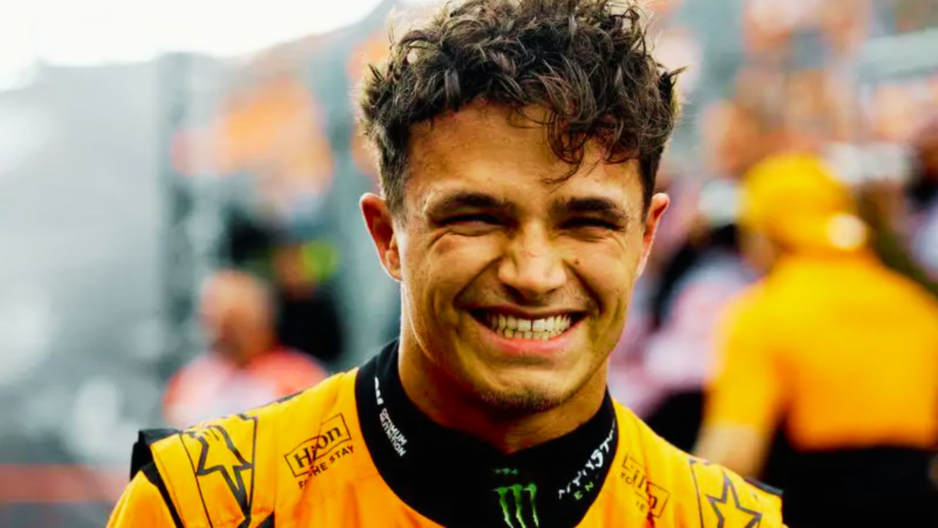 Lando Norris grins in his orange McLaren race suit, hair looking messy and windswept.
