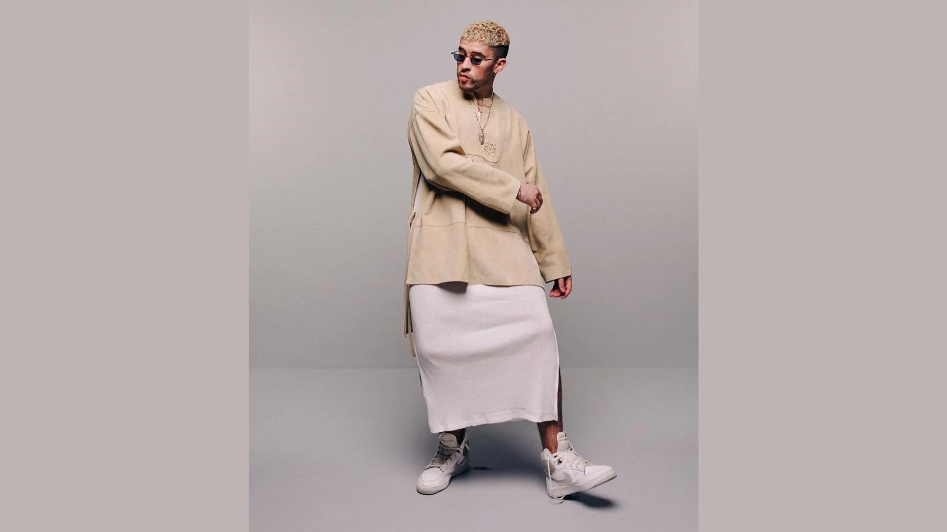 "A person poses in a neutral-toned outfit, wearing a long beige sweater with a white skirt and sneakers. They exude a confident, stylish vibe against a plain background."