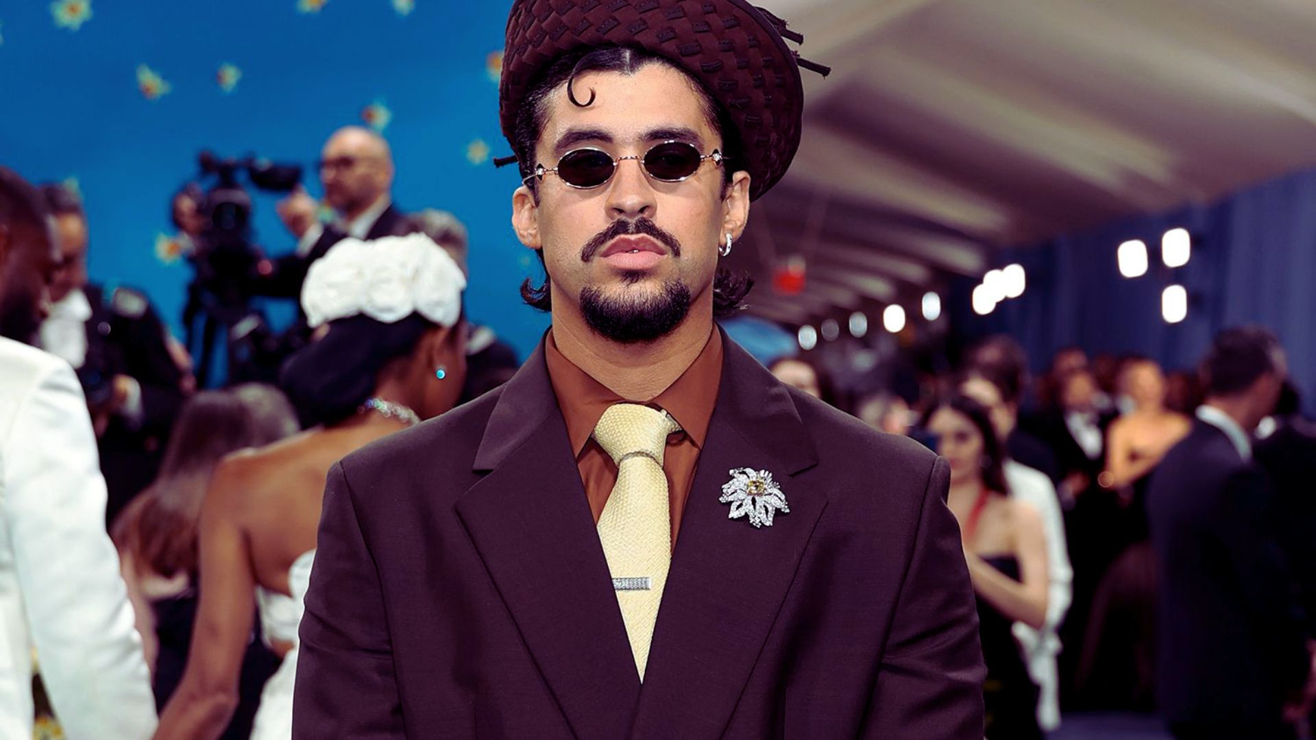 "Man in a brown suit, golden tie, and braided hat poses on a red carpet. He wears sunglasses, and there's a brooch on his lapel.”
