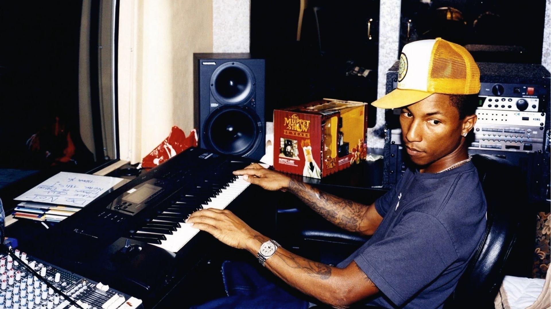 Pharrell Williams at keyboard in studio, yellow trucker hat, navy tee, diamond watch, tattoos