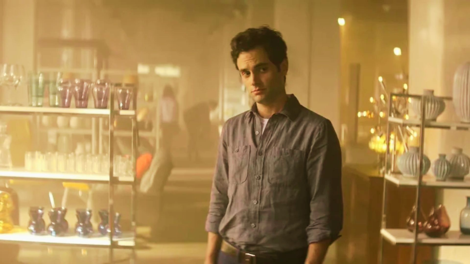 Joe Goldberg is dressed in a rolled-sleeve gray button-up shirt in a warm, golden-lit store.