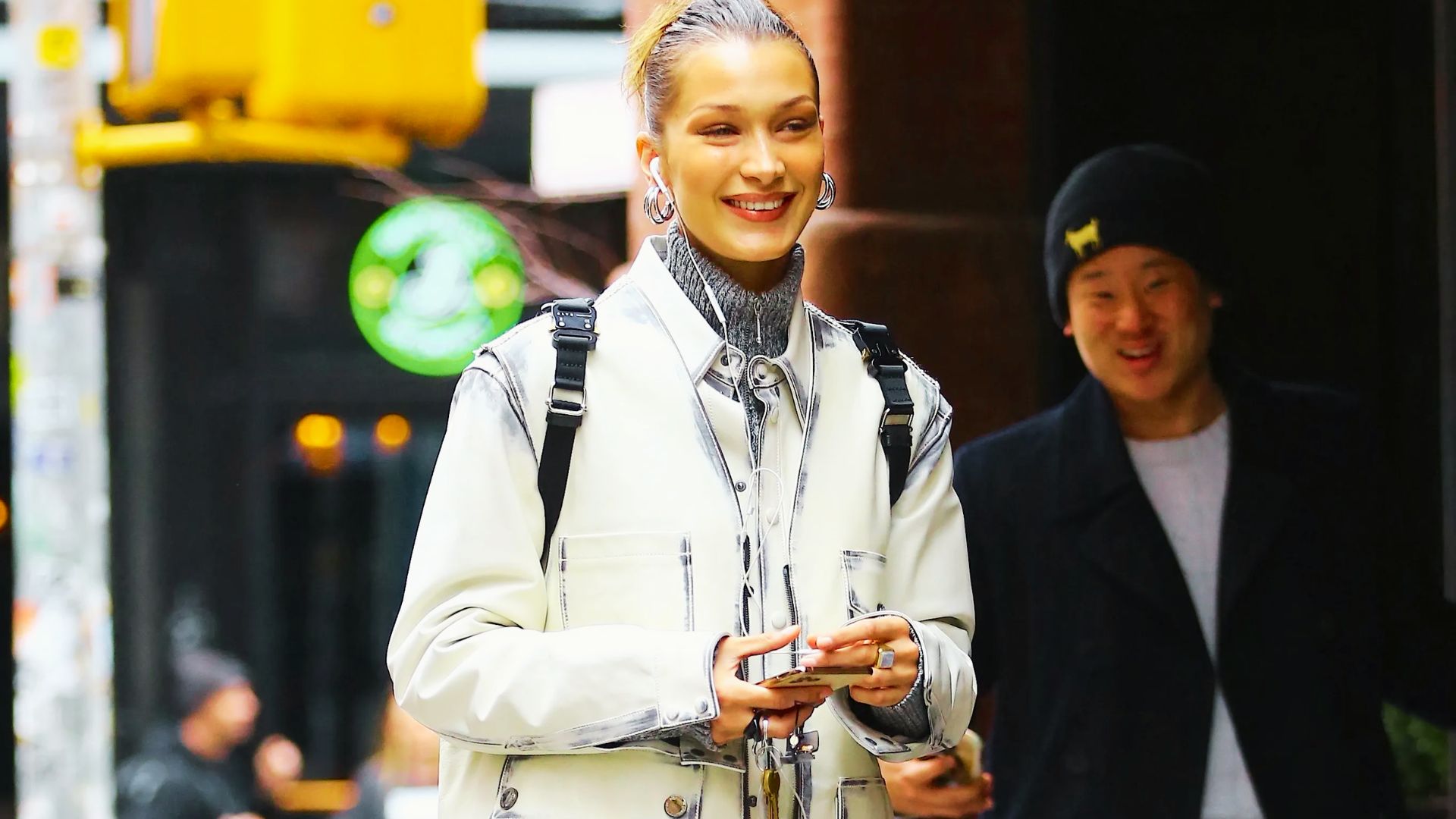 Bella Hadid smiles on a city sidewalk in a white utility jacket layered over a gray turtleneck.