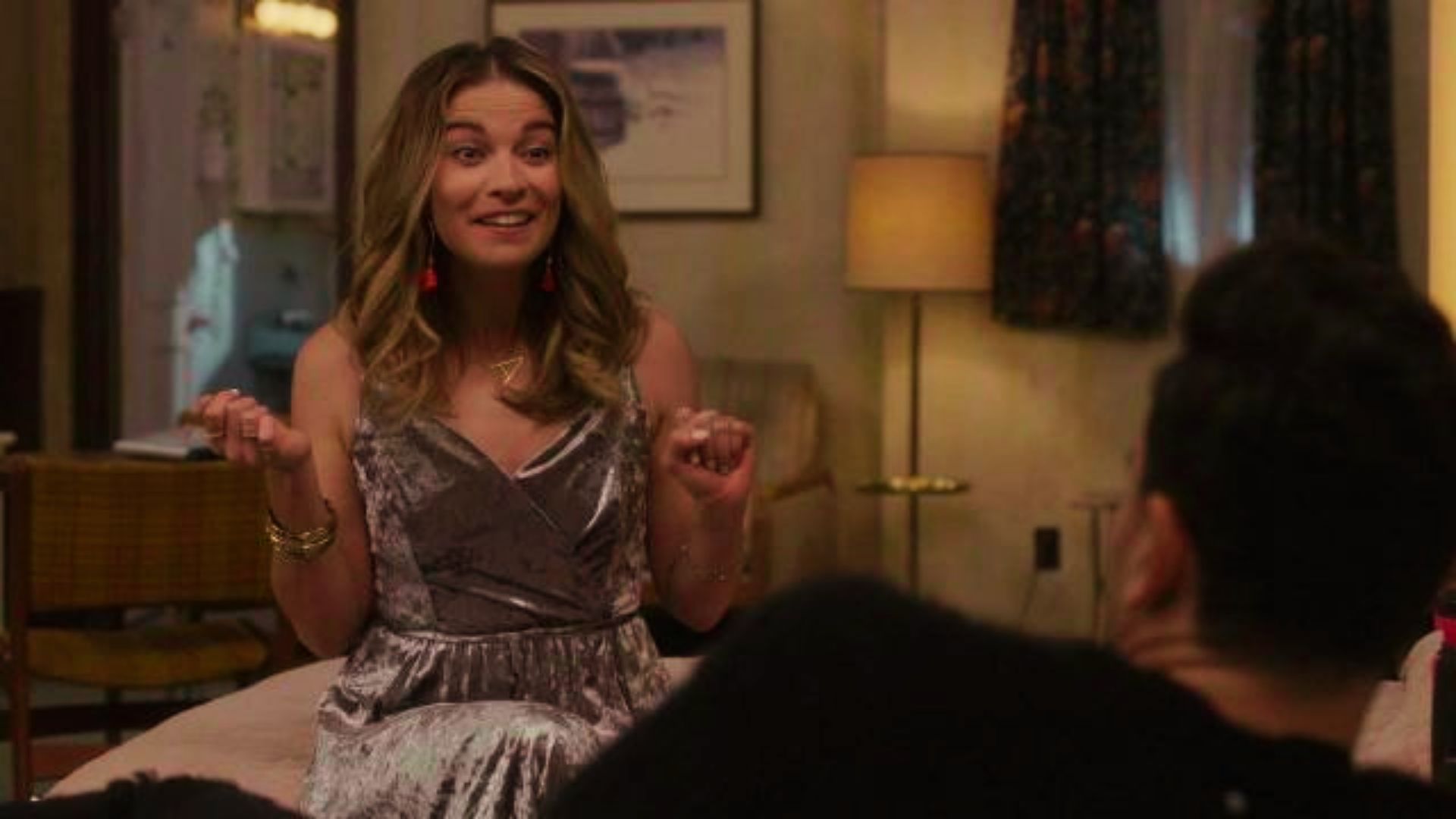 Alexis sits on a motel bed in a silver velvet wrap dress, gesturing while talking.