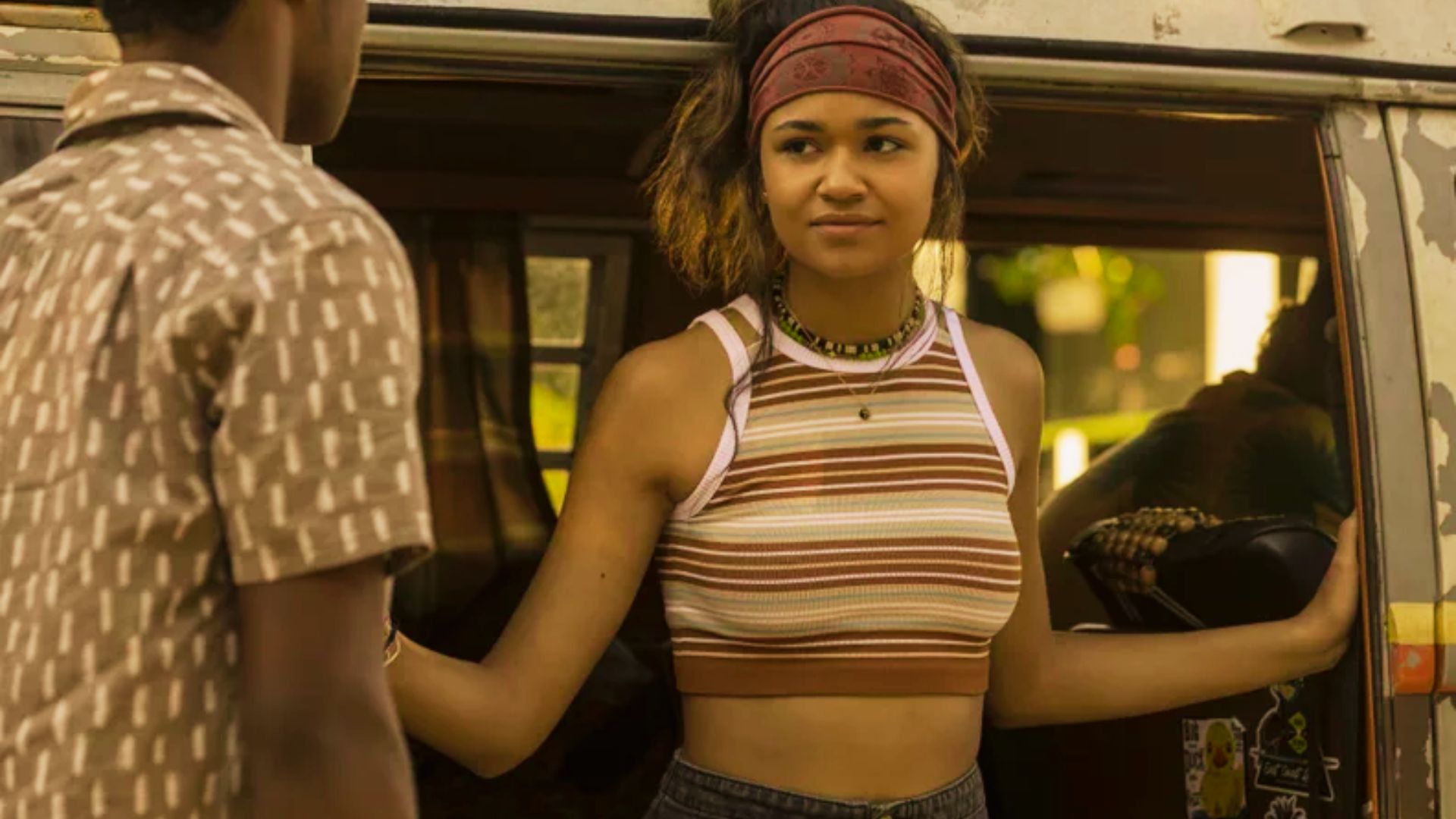 Kiara stands in a van doorway wearing a brown and cream striped halter crop top with a red bandana headband.