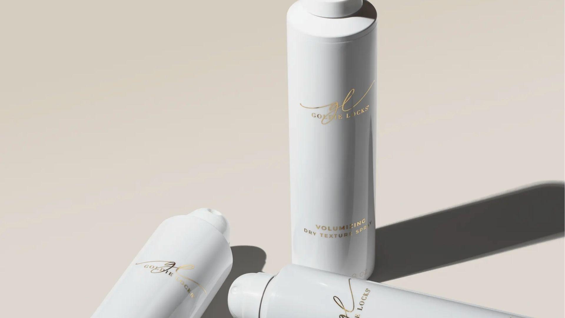 Three white Goldie Locks Volumizing Dry Texture Spray bottles on a beige background