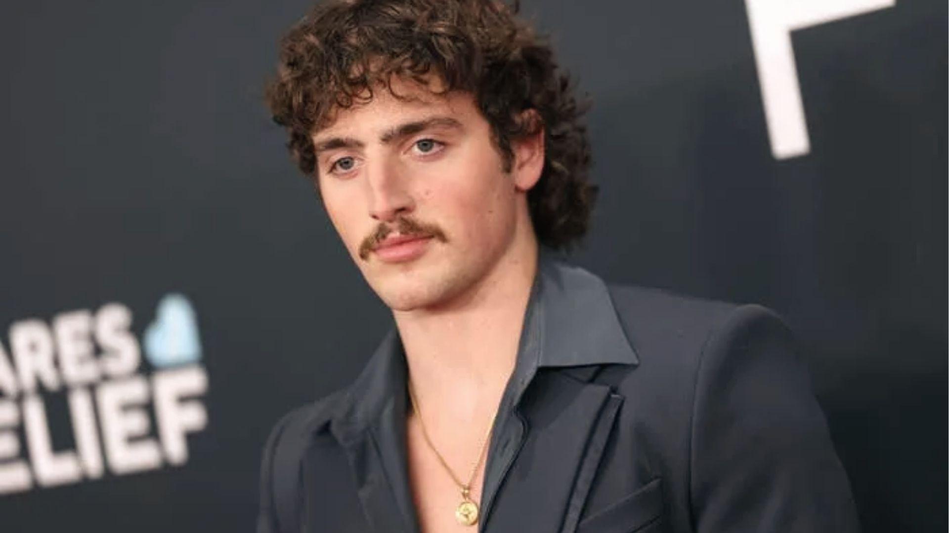 Benson Boone at an event with defined curly brown hair, side part, and mustache.