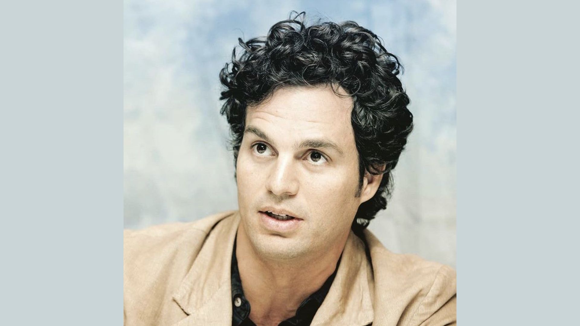 "A man with curly hair wearing a tan jacket stands confidently, showcasing a casual yet stylish appearance."