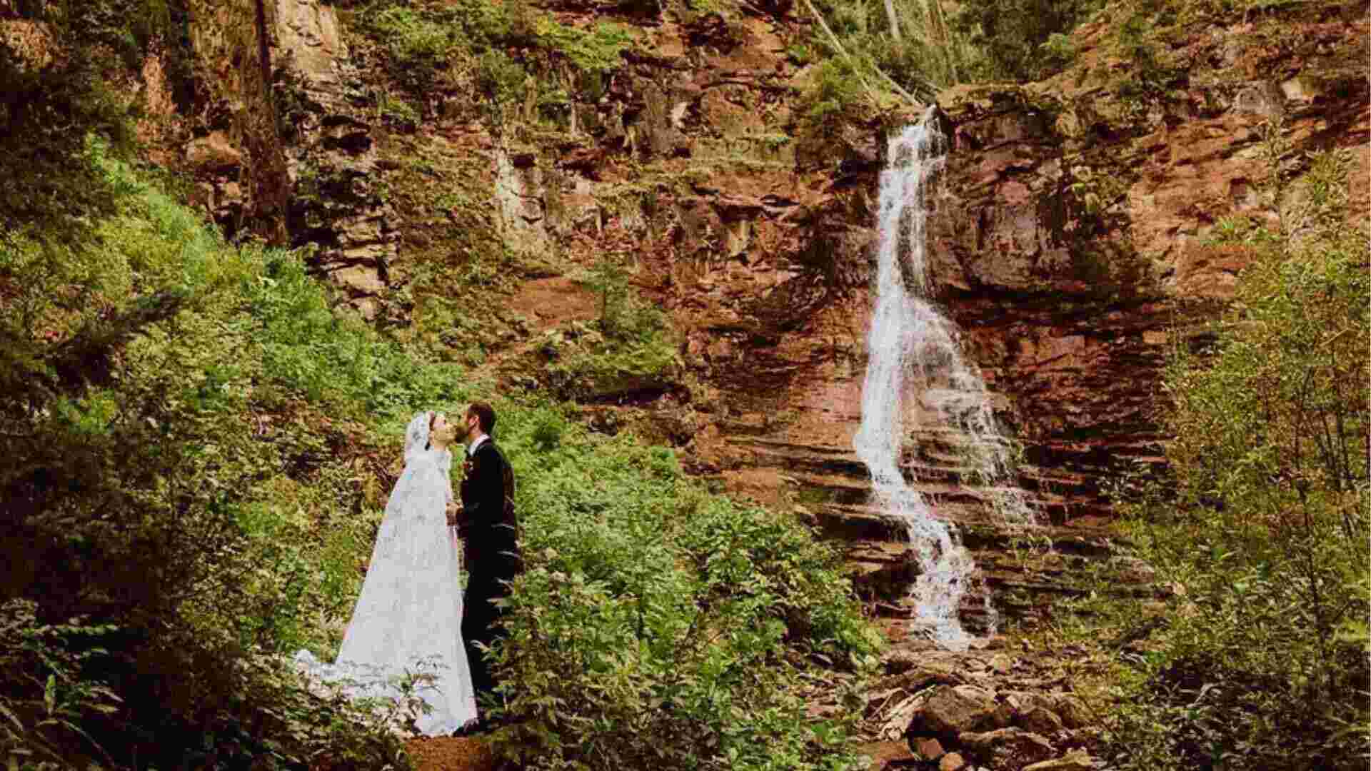 Bride and groom kiss beside waterfall while wearing lace hooded gown and black tuxedo outdoors