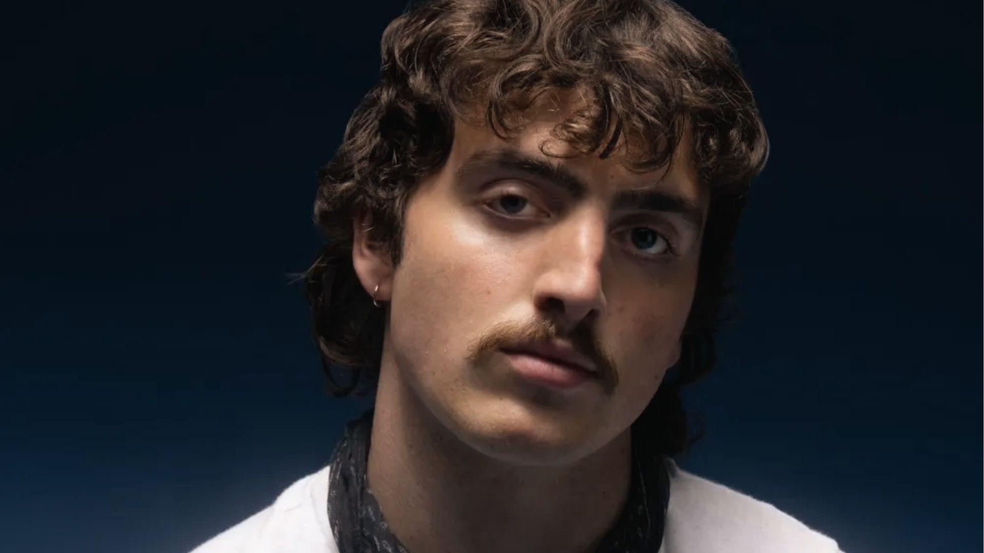 Benson Boone portrait with messy medium curls, face-framing pieces, and mustache.