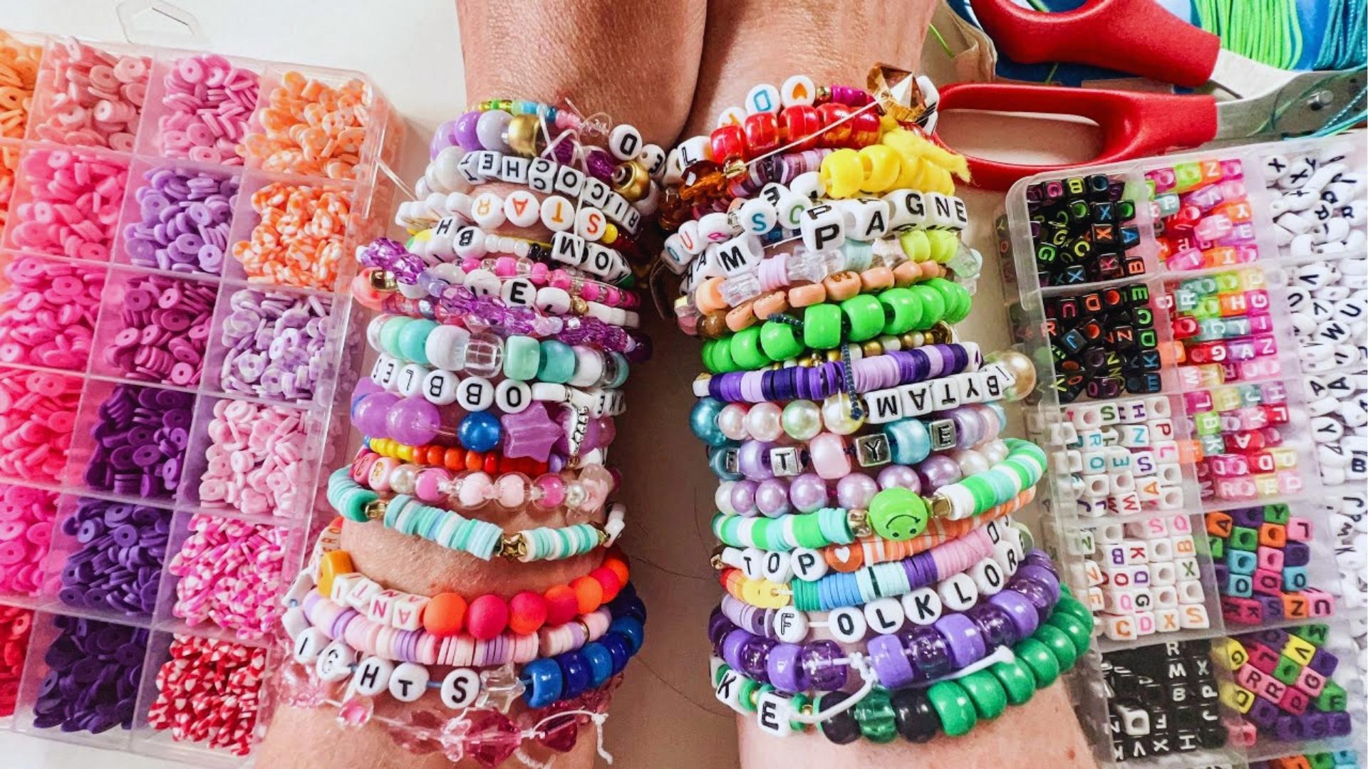 Arms stacked with colorful Taylor Swift bracelets featuring song titles and lyrics, surrounded by bead organizers and crafting supplies