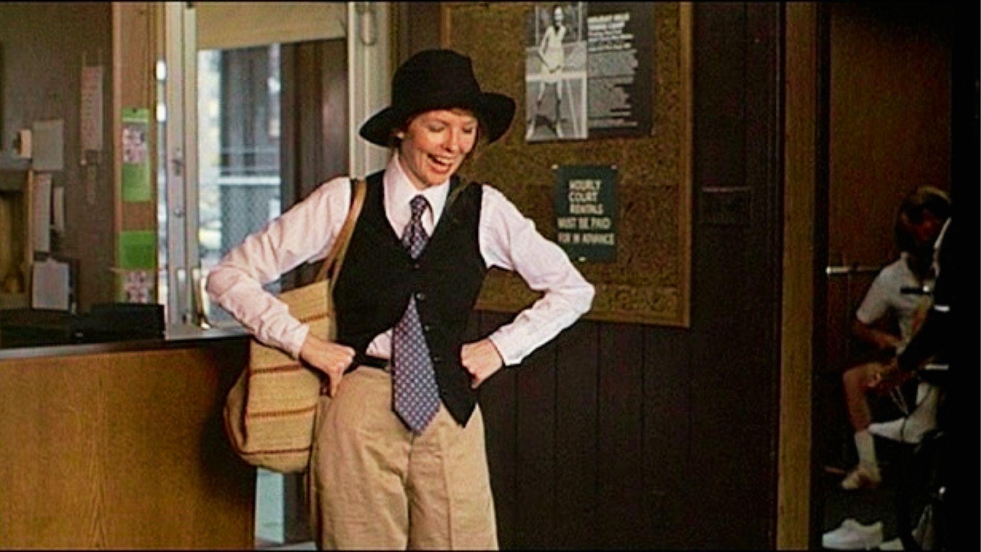 Woman in black vest, white shirt, and tie wears hat at indoor film set