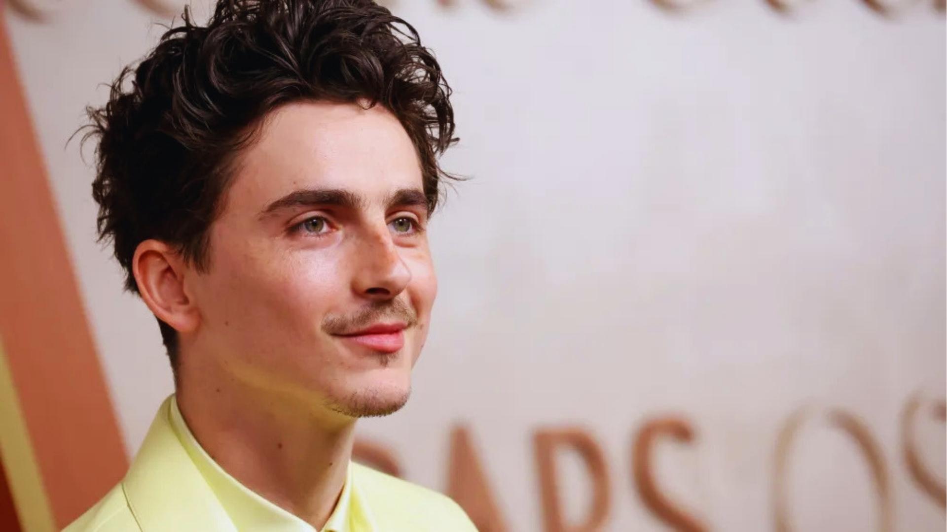 Timothée Chalamet before buzz cut with tousled dark curls returning, pairing a yellow suit with light facial hair.