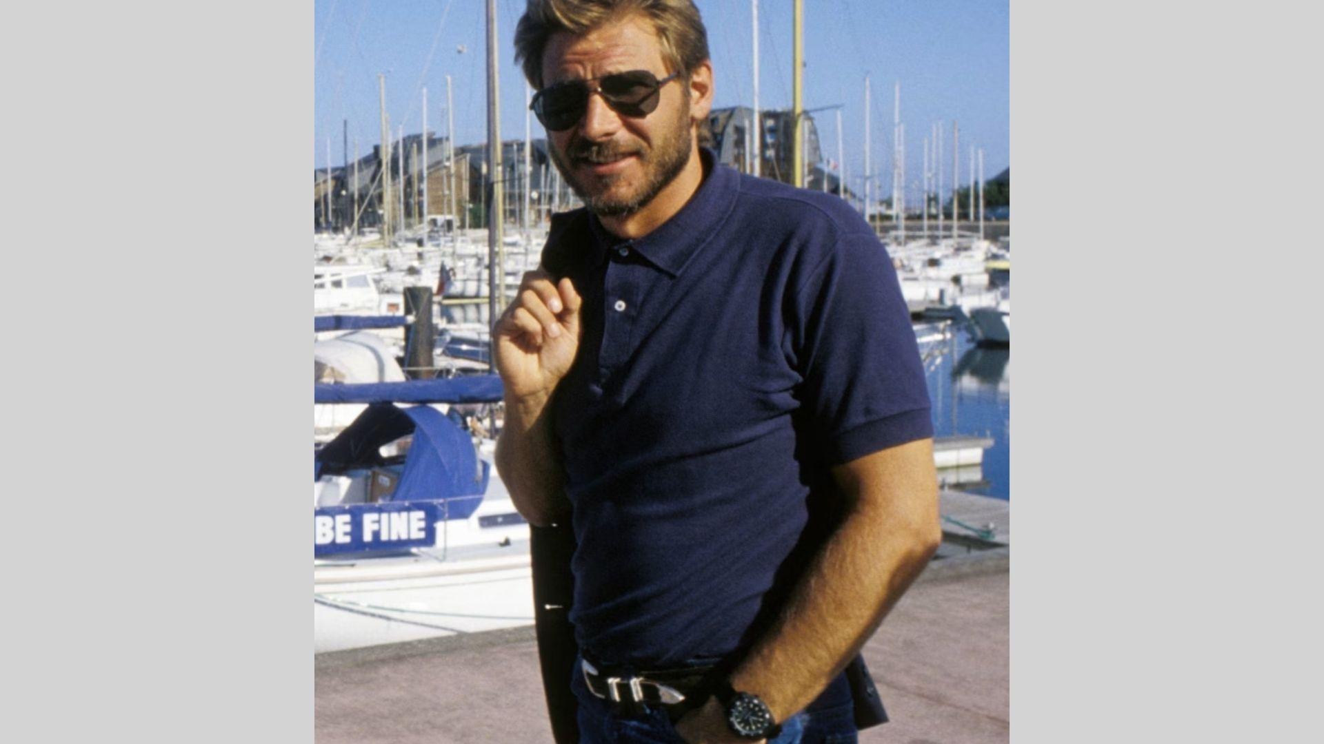Harrison Ford stands at marina in navy polo and sunglasses holding jacket over shoulder