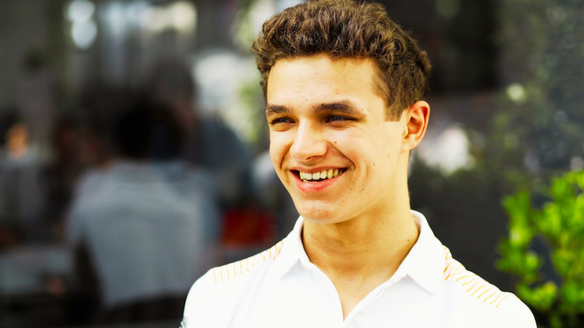 Lando Norris smiles in a white McLaren polo, showing a neater, more styled curly haircut.