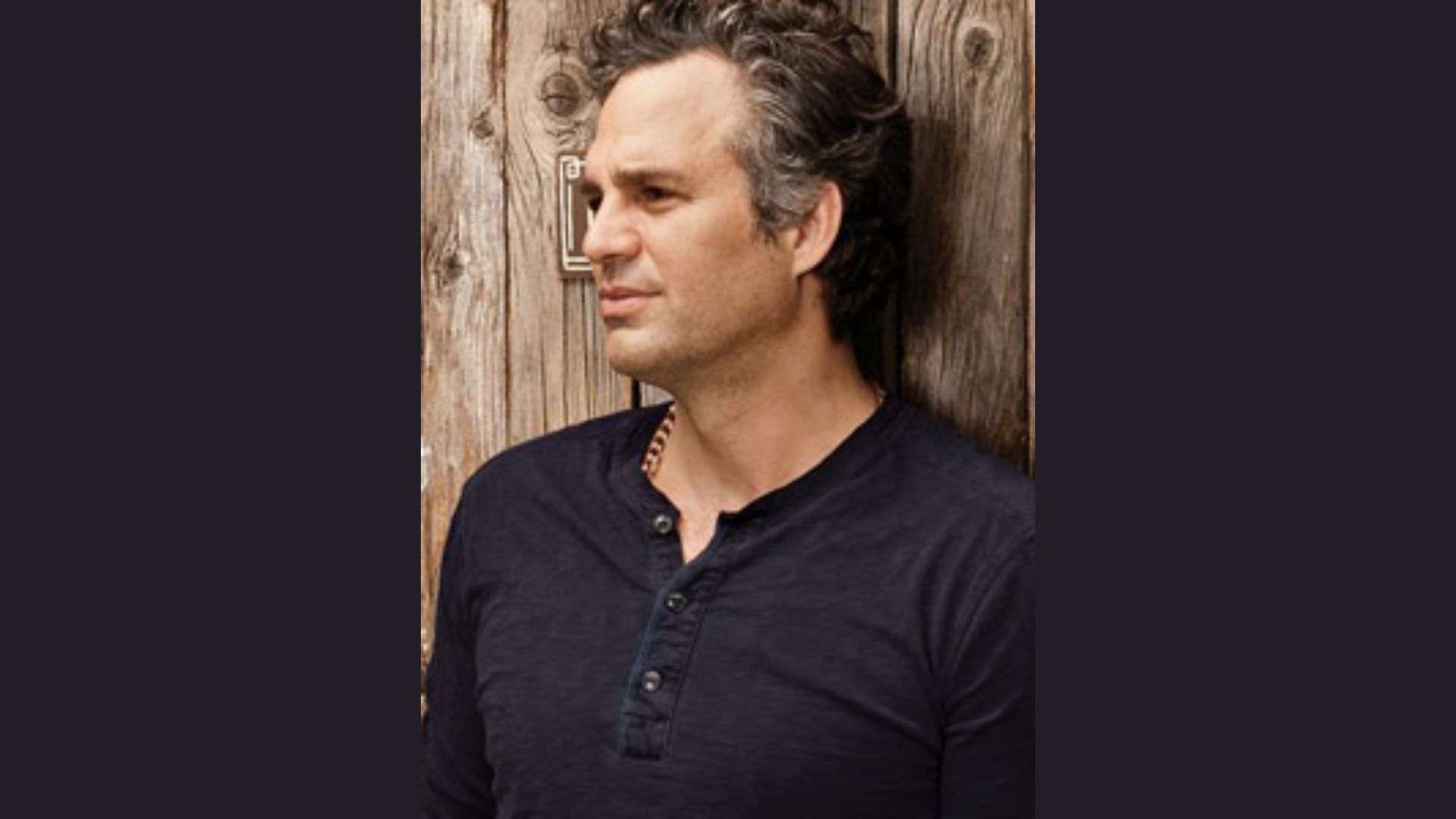 "A man with curly hair in a black shirt leans against a wooden wall, creating a relaxed and casual atmosphere."