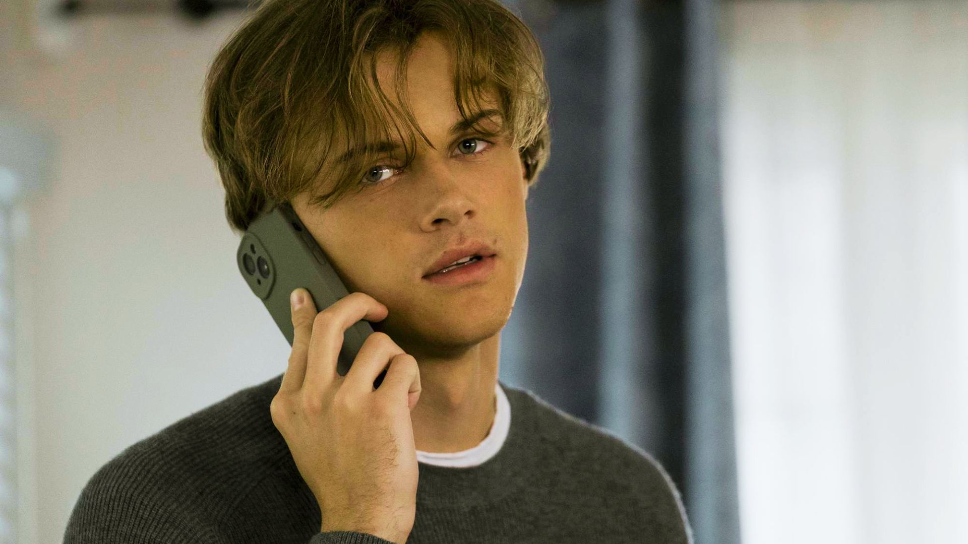 Conrad Fisher on a phone call, wearing a charcoal ribbed crewneck sweater layered over a white tee.