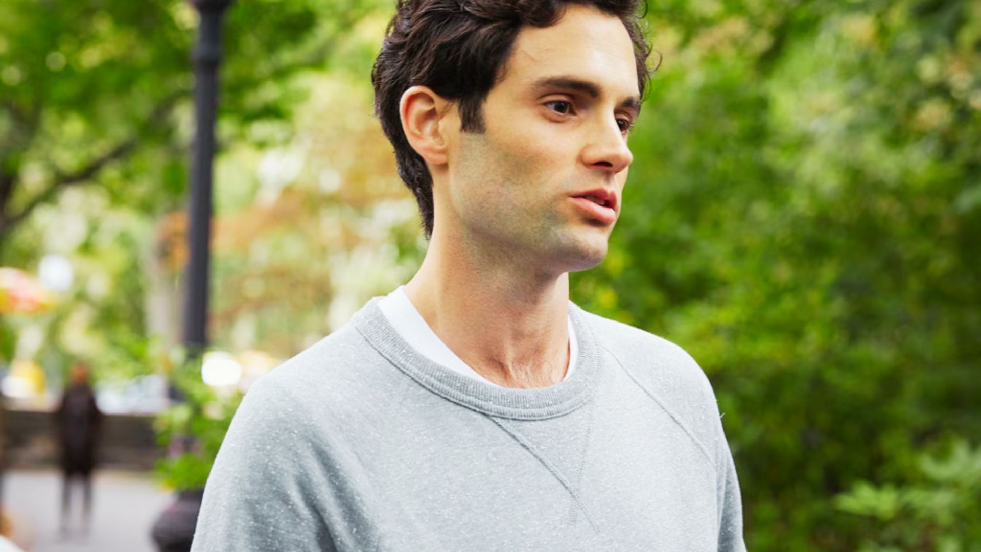Joe Goldberg wears a light gray crewneck sweatshirt layered over a white tee outdoors.
