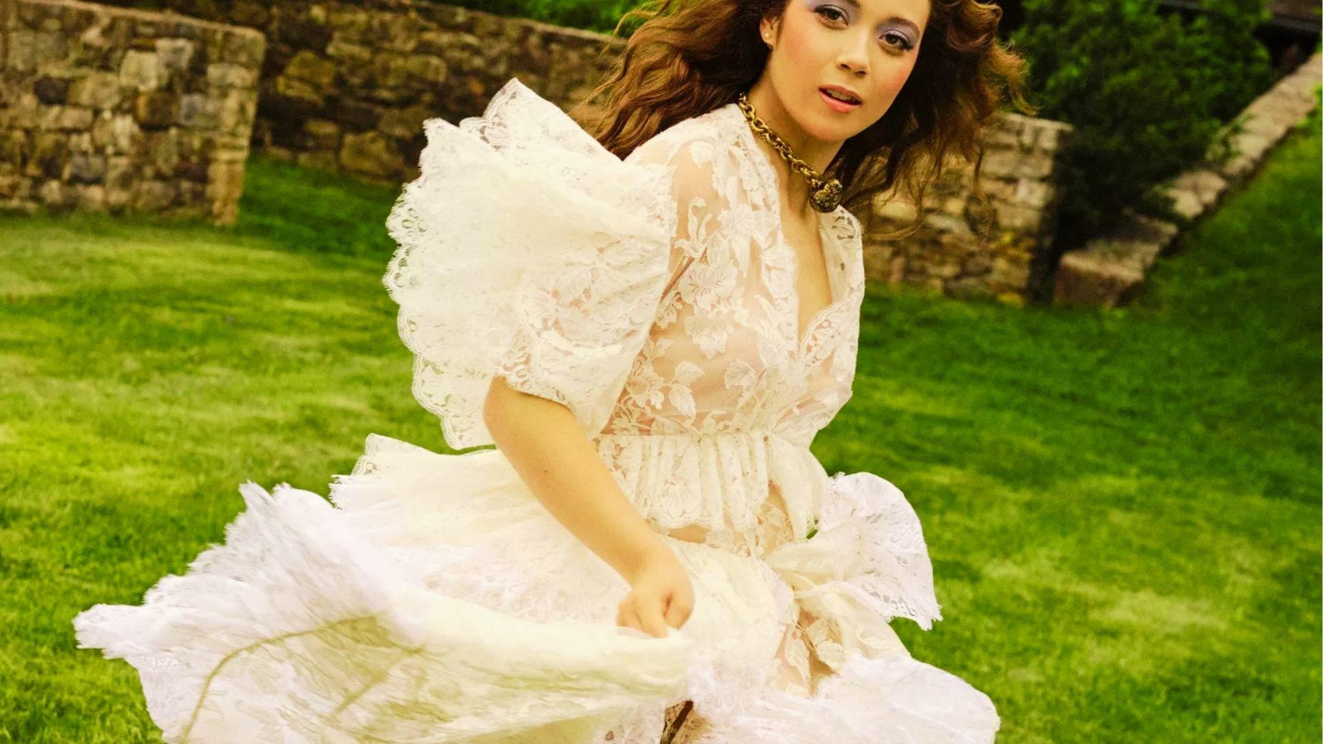 Woman twirls in white lace dress with puff sleeves and gold necklace on green grass