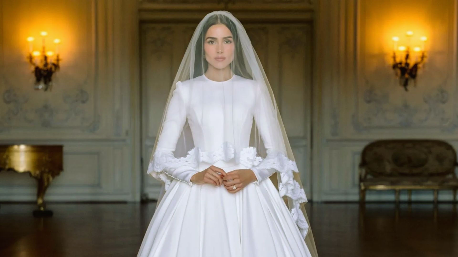 "Bride in a classic white gown stands in an ornate room with soft lighting. Her veil cascades down, and her expression is serene."