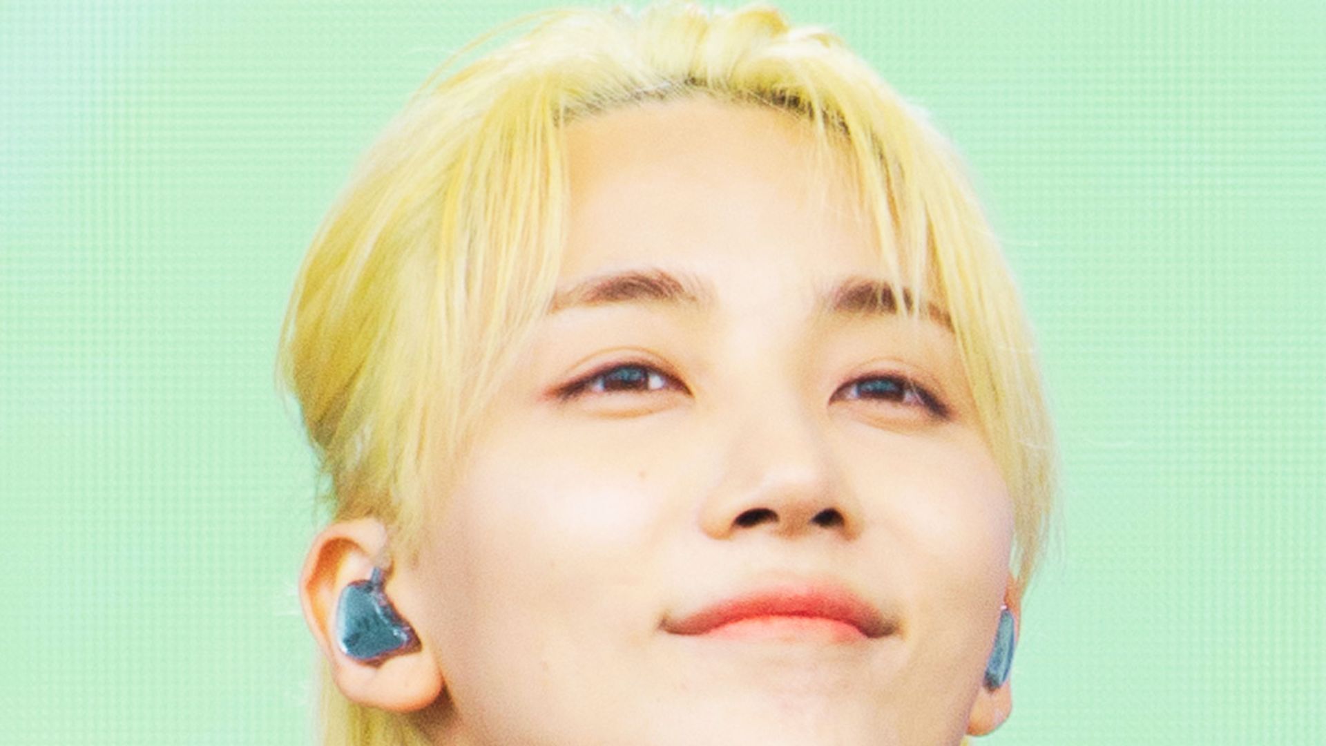 Stage shot of Jeonghan smiling with slicked-back platinum blonde hair and in-ear monitors.