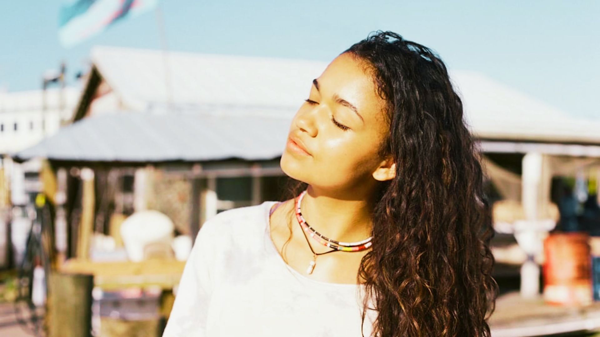 Kiara soaks up the sun at a marina in a loose white top, layered beaded necklaces, and wet curly hair.