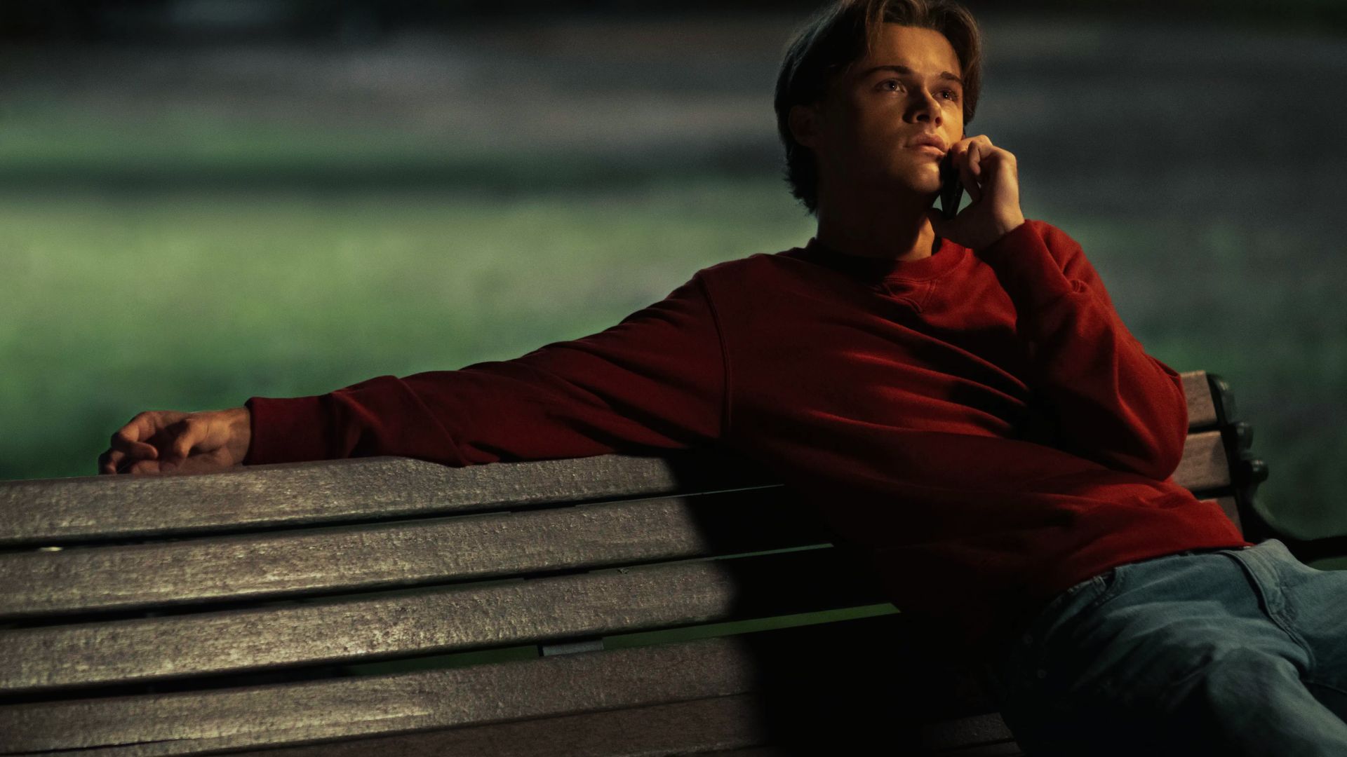 Conrad leans on a bench at night in a deep red sweatshirt and jeans while on a call.