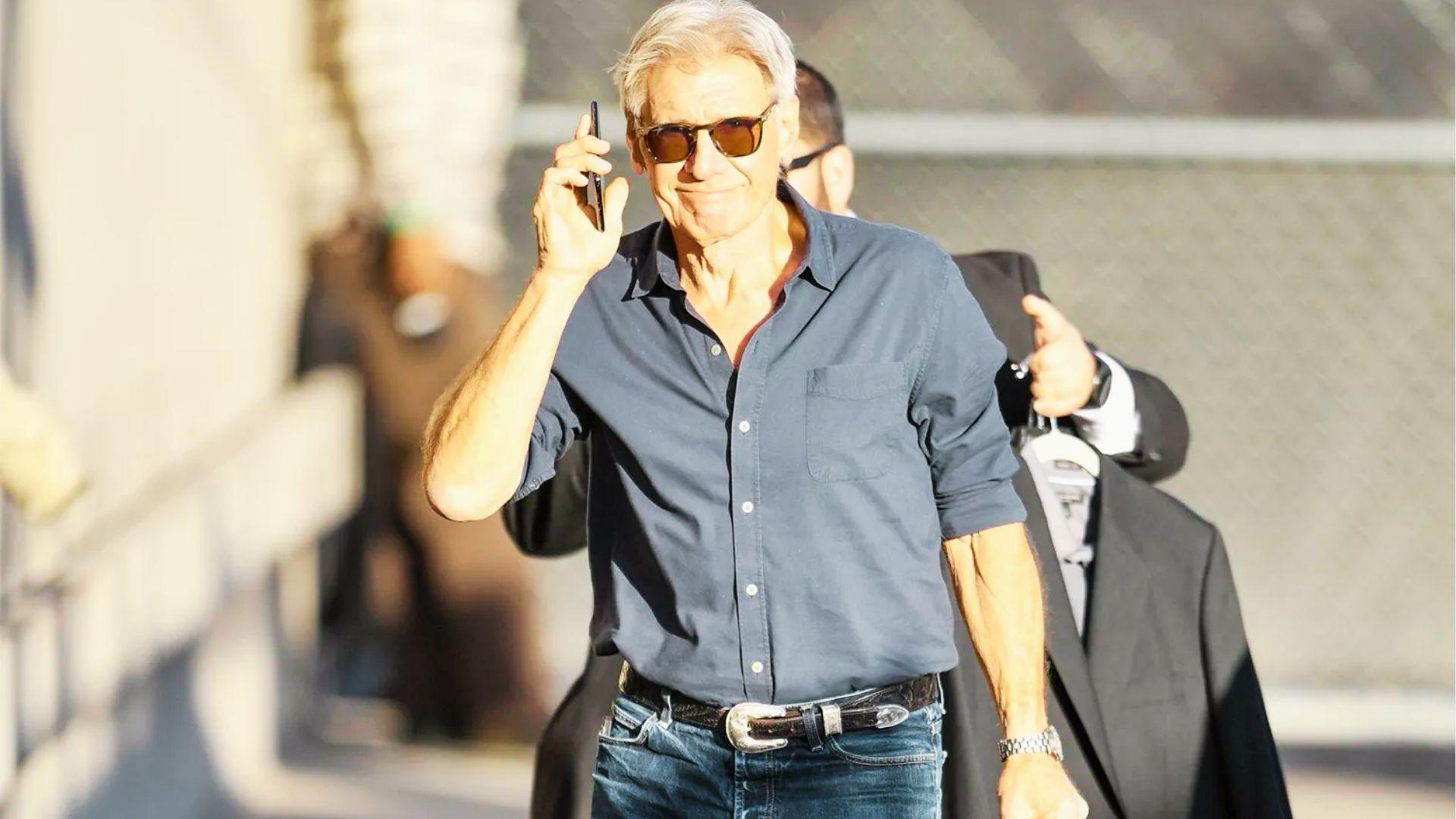 Older Harrison Ford walks smiling in blue shirt and jeans while talking on phone outdoors