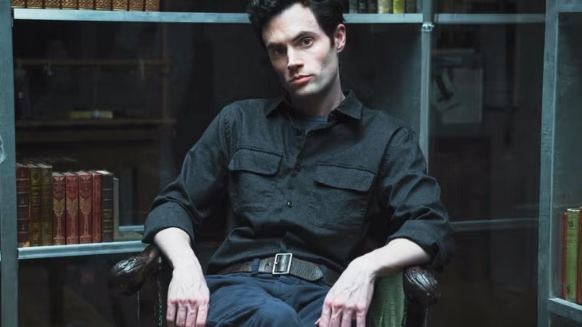 Joe Goldberg sits in a dimly lit library wearing a black work shirt with chest pockets.