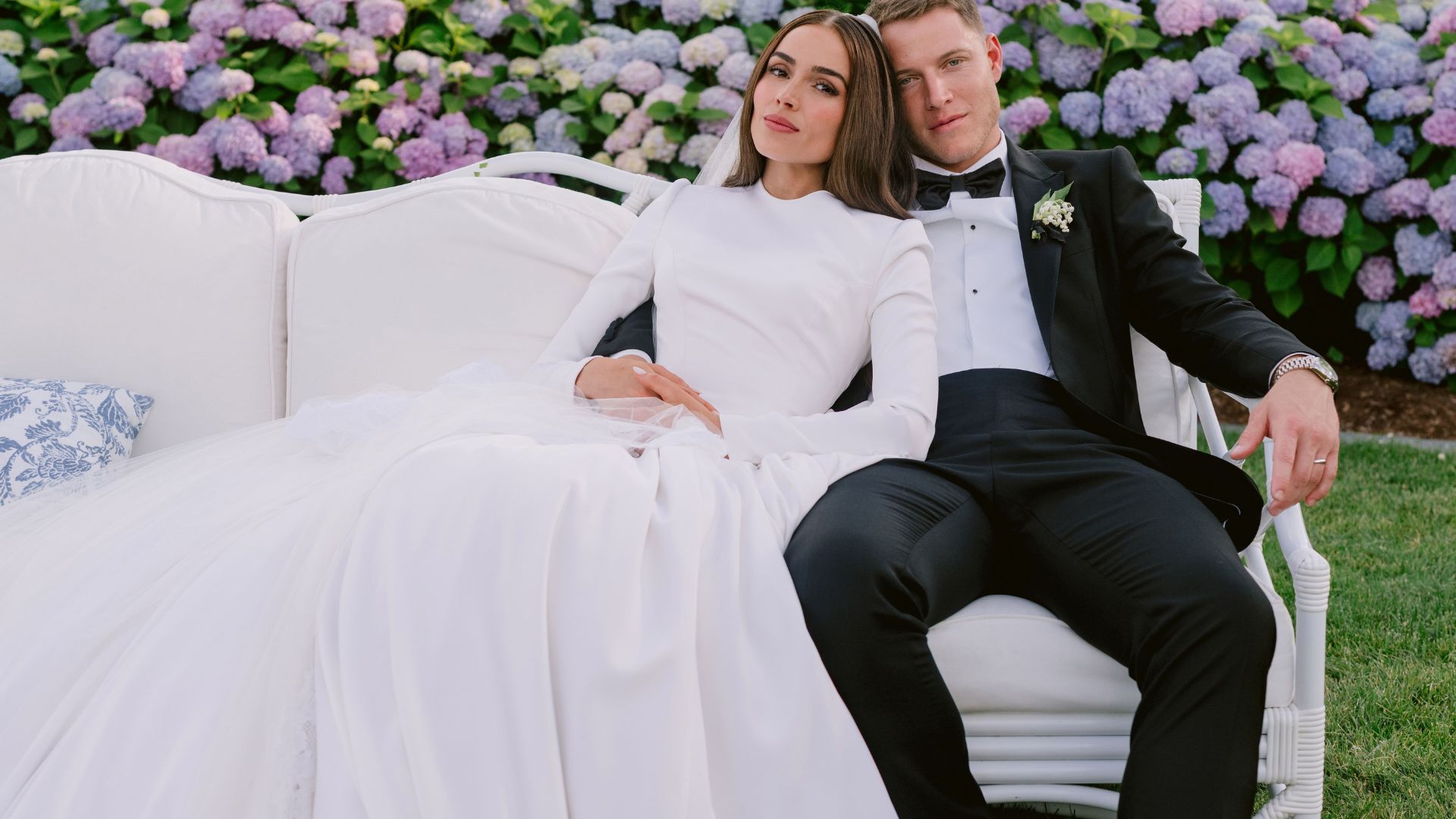 "A couple in wedding attire sits on a white couch outdoors, against a backdrop of blooming hydrangeas. The bride wears a long white gown, and the groom is in a black tuxedo. They appear relaxed and content."