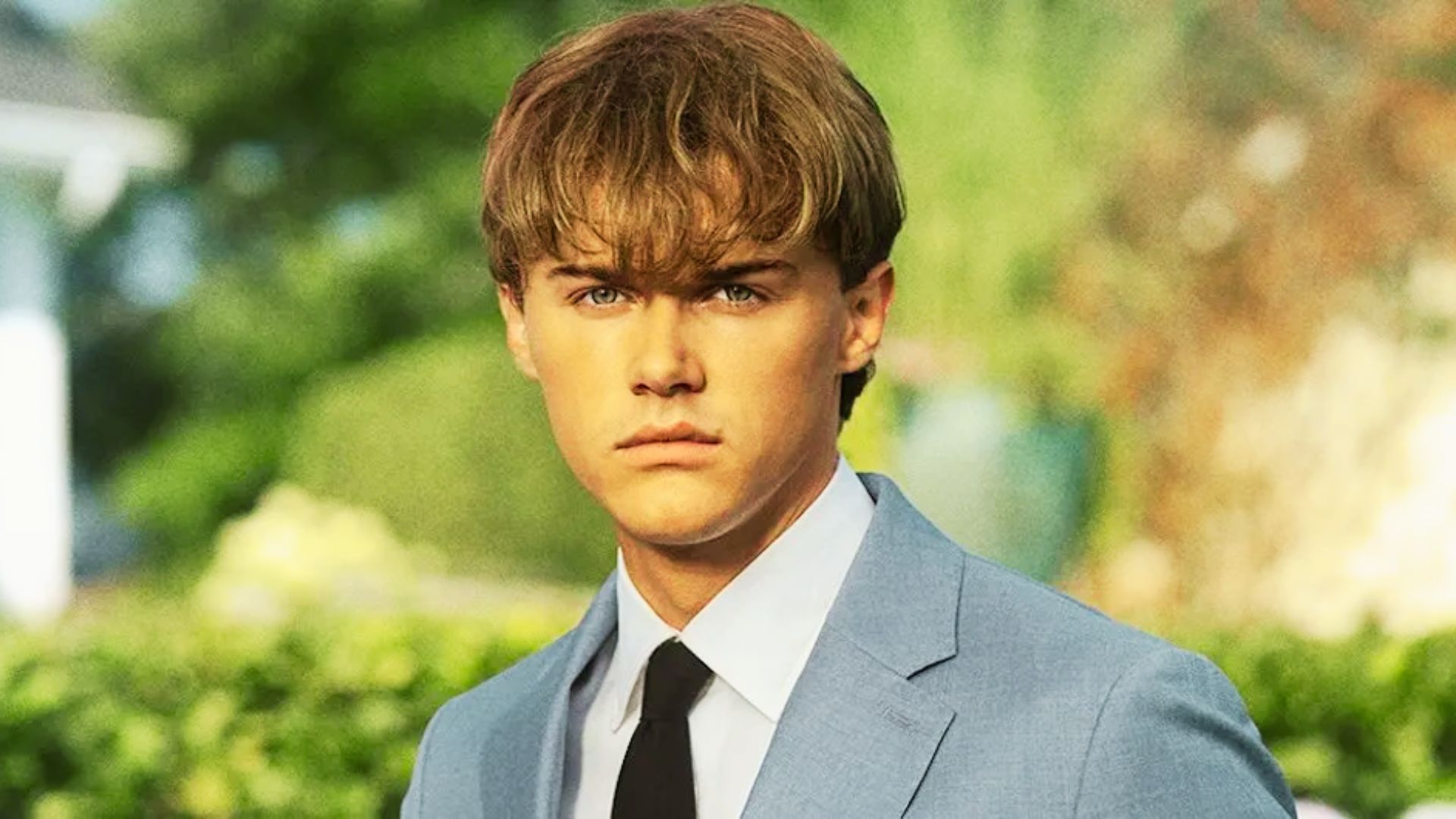 Conrad stands outdoors in a light blue suit with a white shirt and black tie.