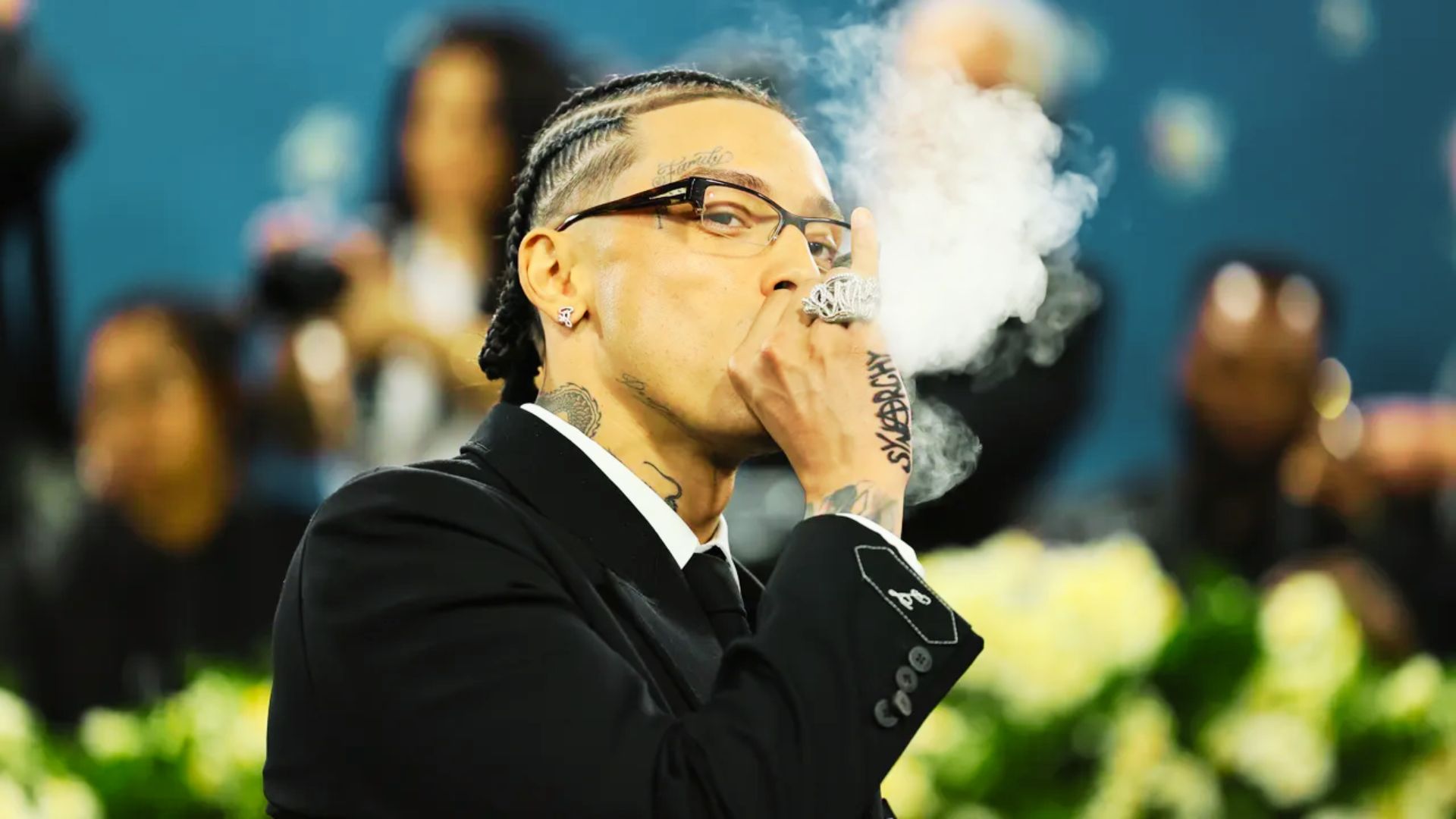 Central Cee exhales smoke in a black suit and glasses, wearing tight cornrows braided back with a sharp hairline.